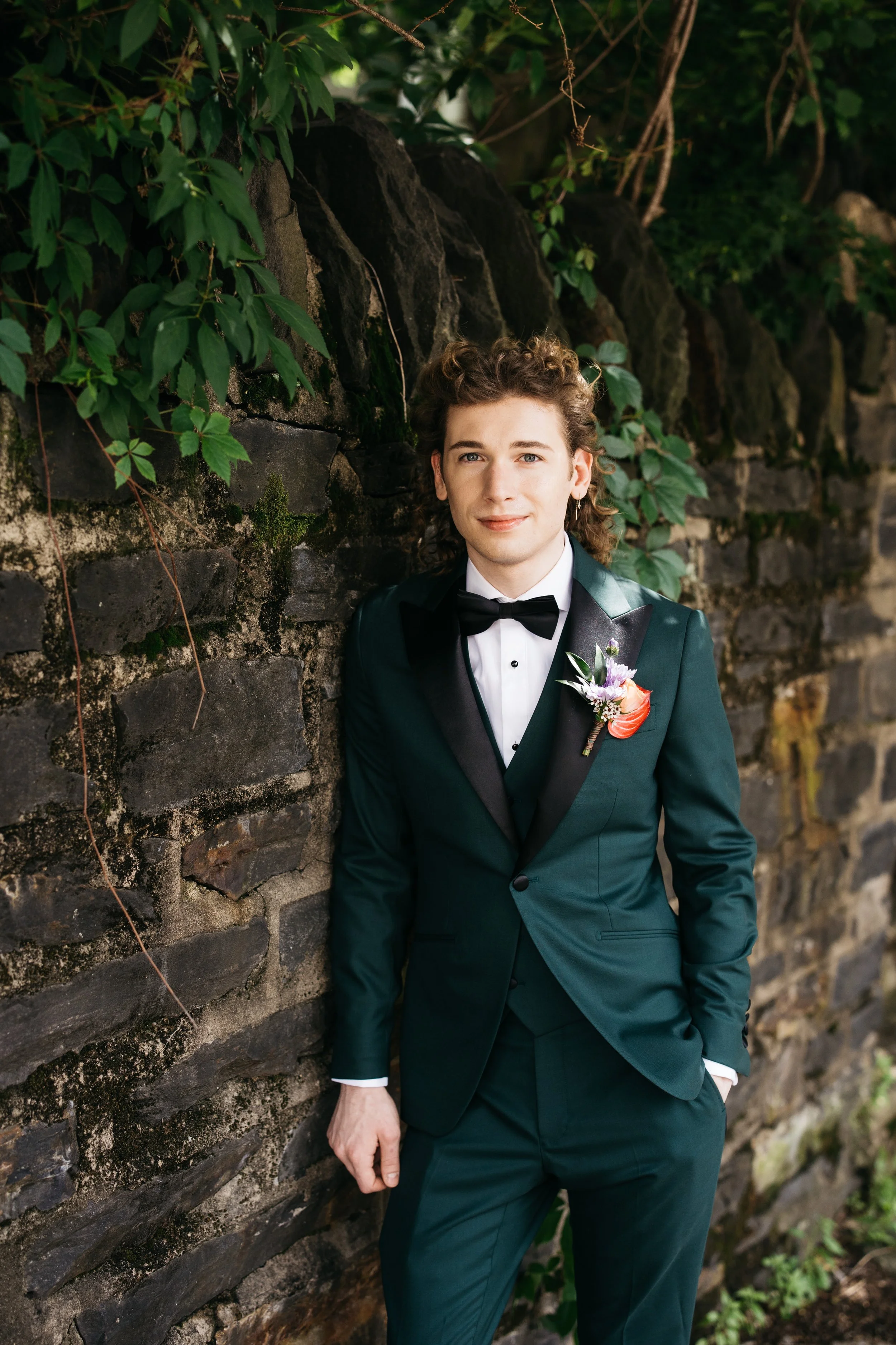 A young man in a tuxedo with a boutonniere stands against a stone and ivy wall.