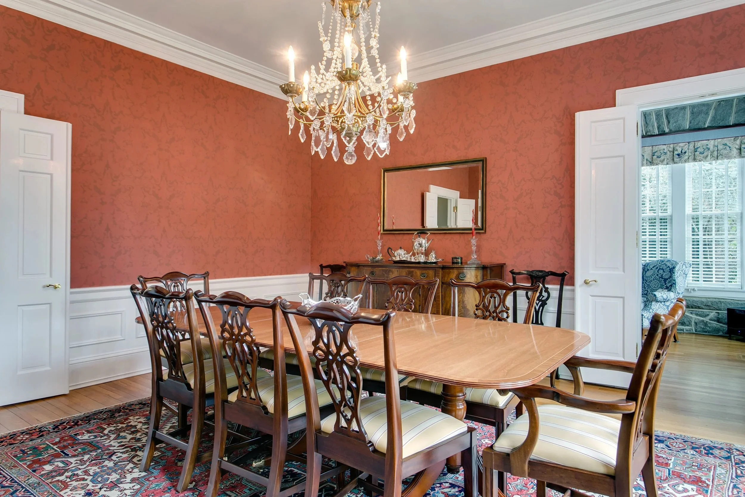 A dining room with a large wooden table surrounded by ten dark wooden chairs with striped upholstery. The room has reddish-orange wallpaper, white crown molding, and white wainscoting. A crystal chandelier hangs from the ceiling, and a wooden sideboa