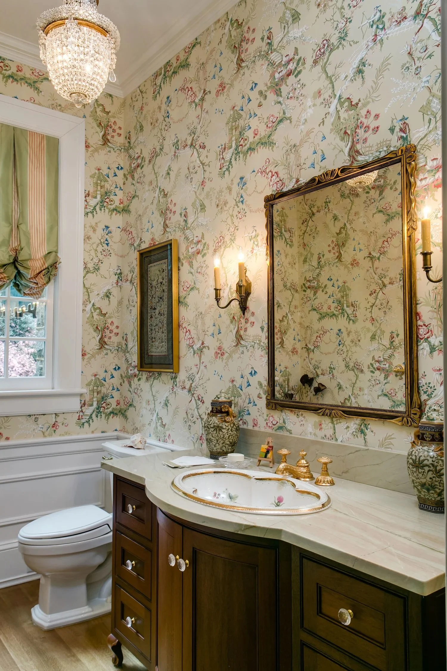 A vintage-style bathroom with floral wallpaper, a large mirror with a gold frame, a marble countertop with a decorative sink, gold fixtures, a large vase, wall sconces, a window with curtains, and a chandelier ceiling light.