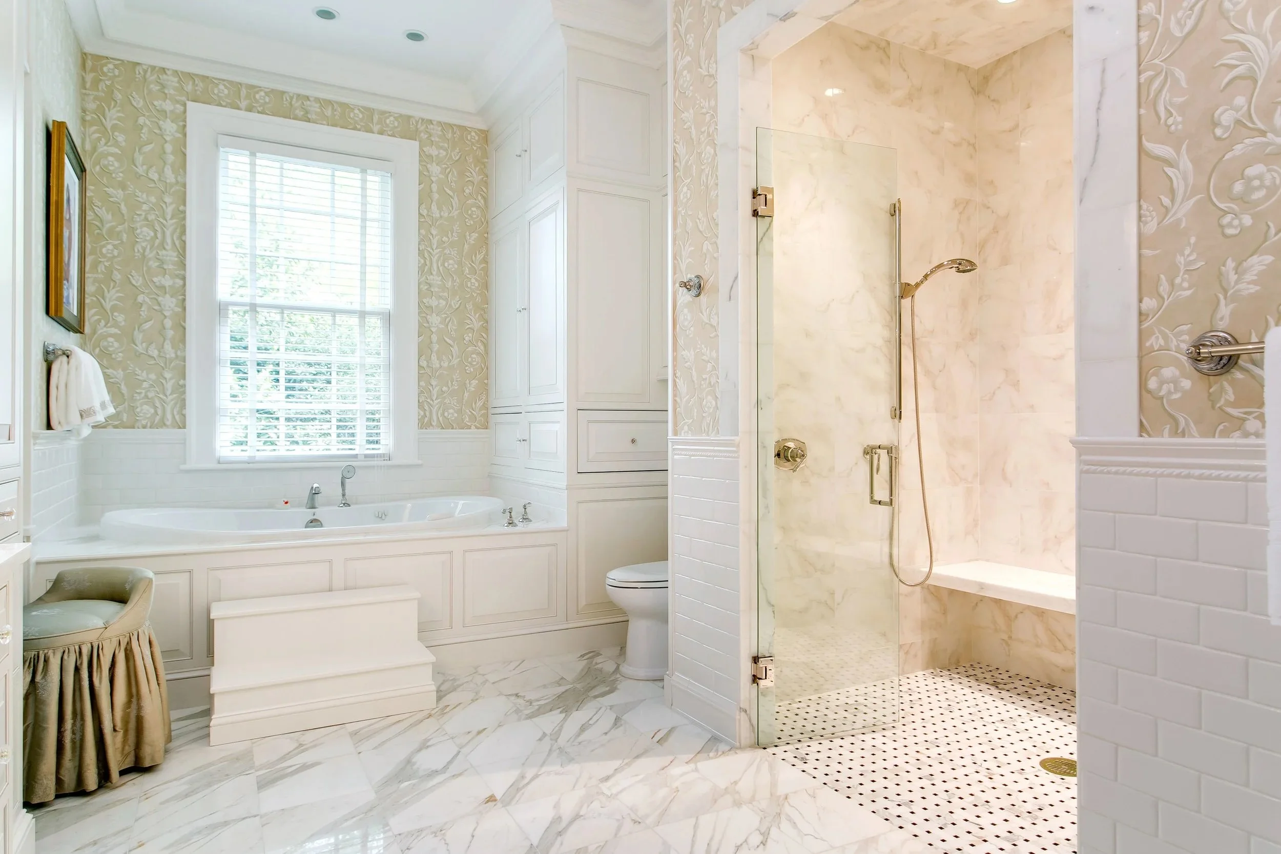 A luxurious bathroom featuring a large built-in bathtub with ornate white cabinetry, a marble shower with a glass door and gold fixtures, and a window with white blinds, all decorated with elegant floral wallpaper and white wainscoting.