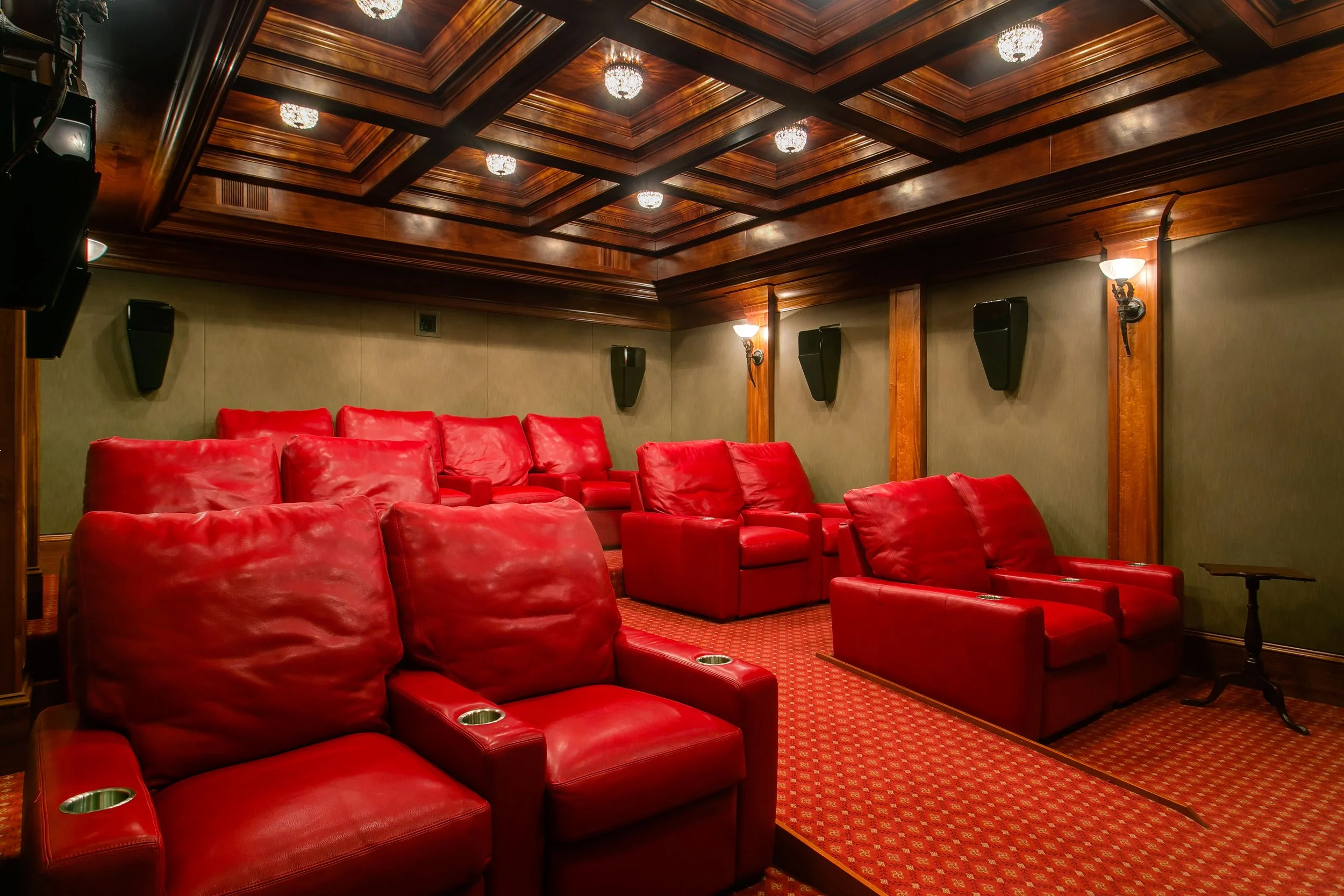 A cozy private movie theater room with red leather seats, dark wood paneling, a patterned red carpet, and large black speakers on the walls.