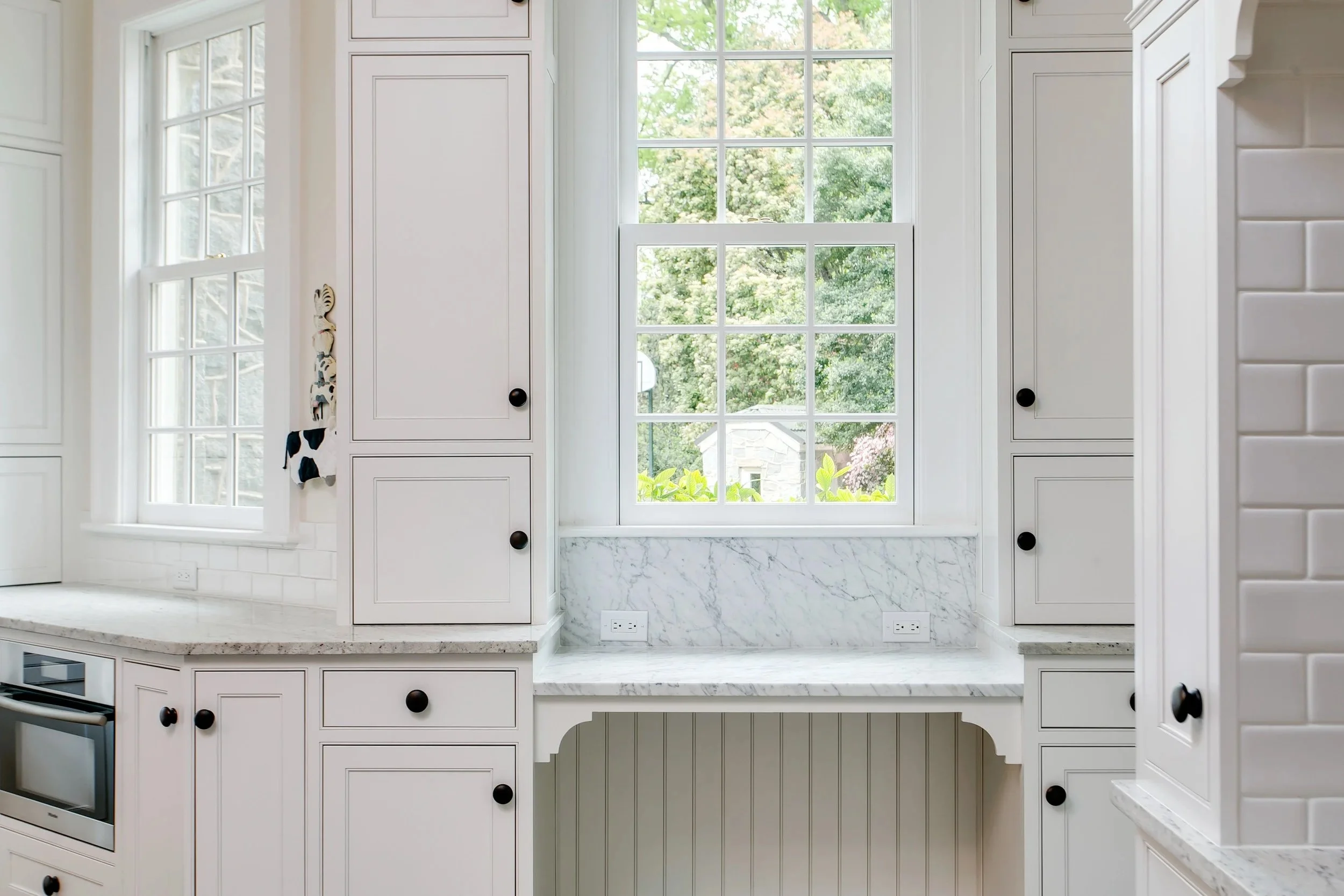 White kitchen window with a view of trees outside, marble countertop below, and white cabinets with black knobs.
