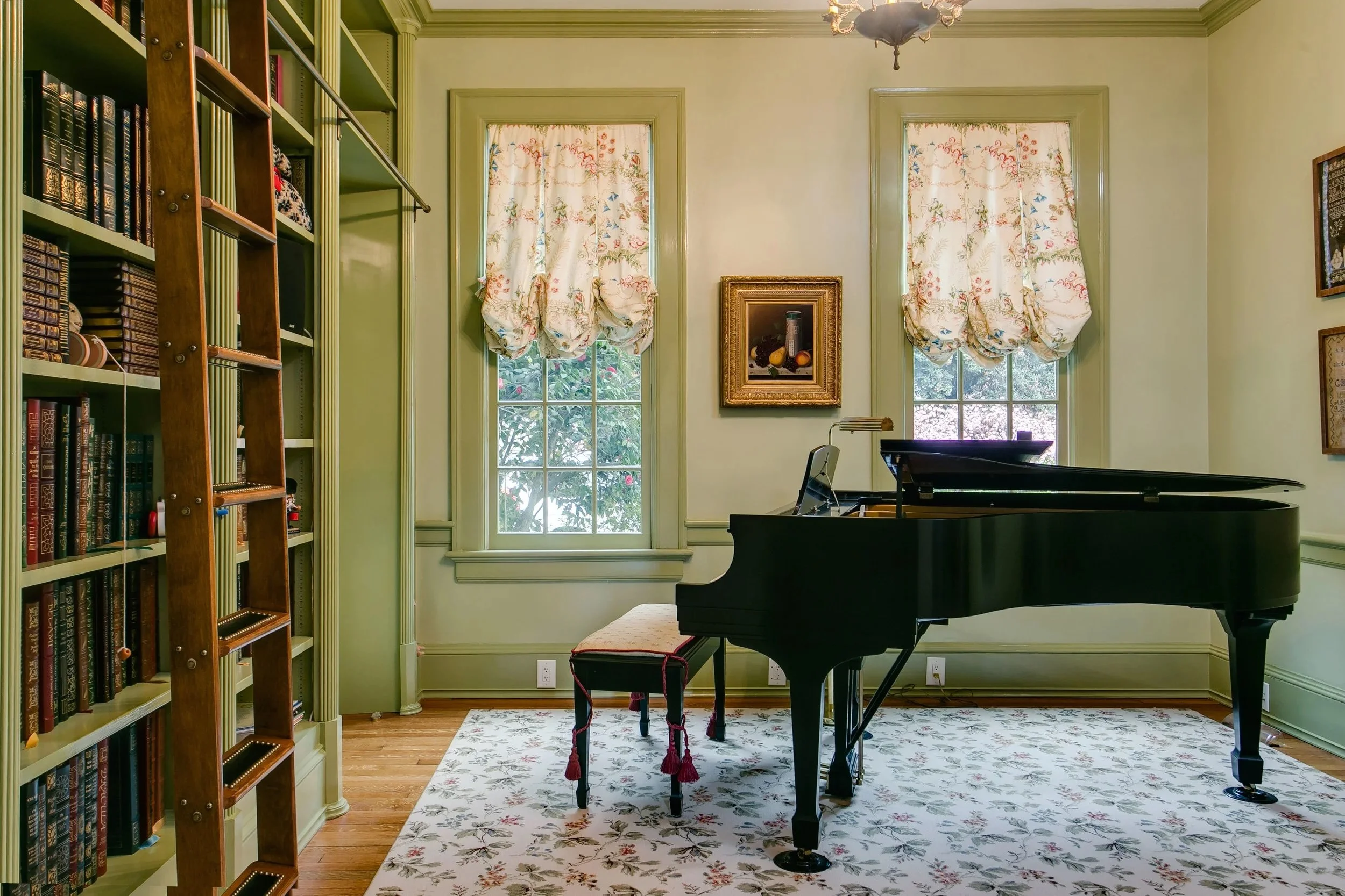 A room with green walls and trim, two large windows with floral curtains, a grand black piano with a matching bench, a framed still life picture on the wall, and a patterned rug on the wooden floor. There is a chandelier hanging from the ceiling.