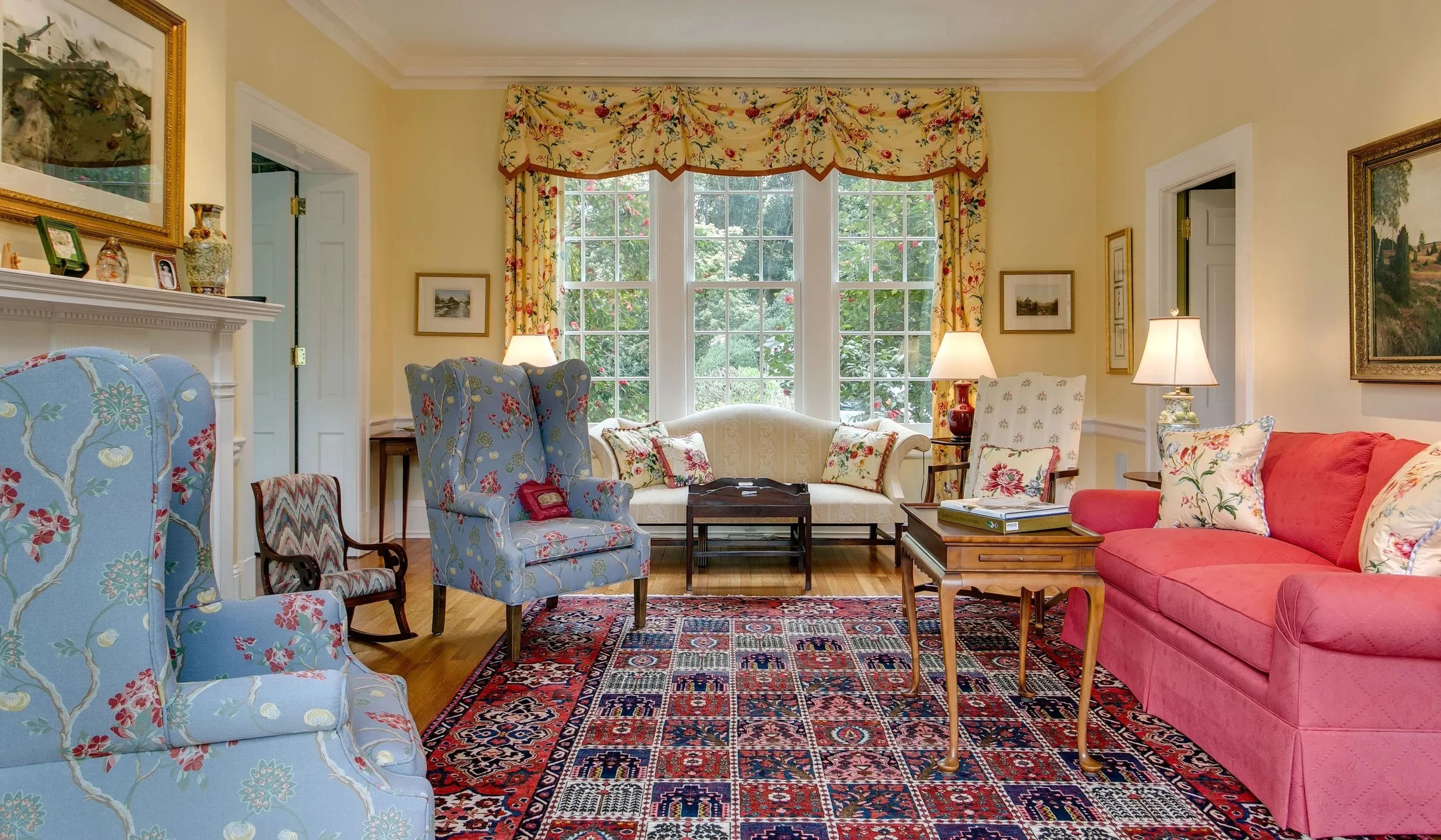 A cozy living room with yellow walls, large bay window with floral curtains, and vintage furniture. There are blue, white, and pink armchairs, a red sofa, and a Persian rug on the hardwood floor. The room is decorated with artwork, lamps, and decorat