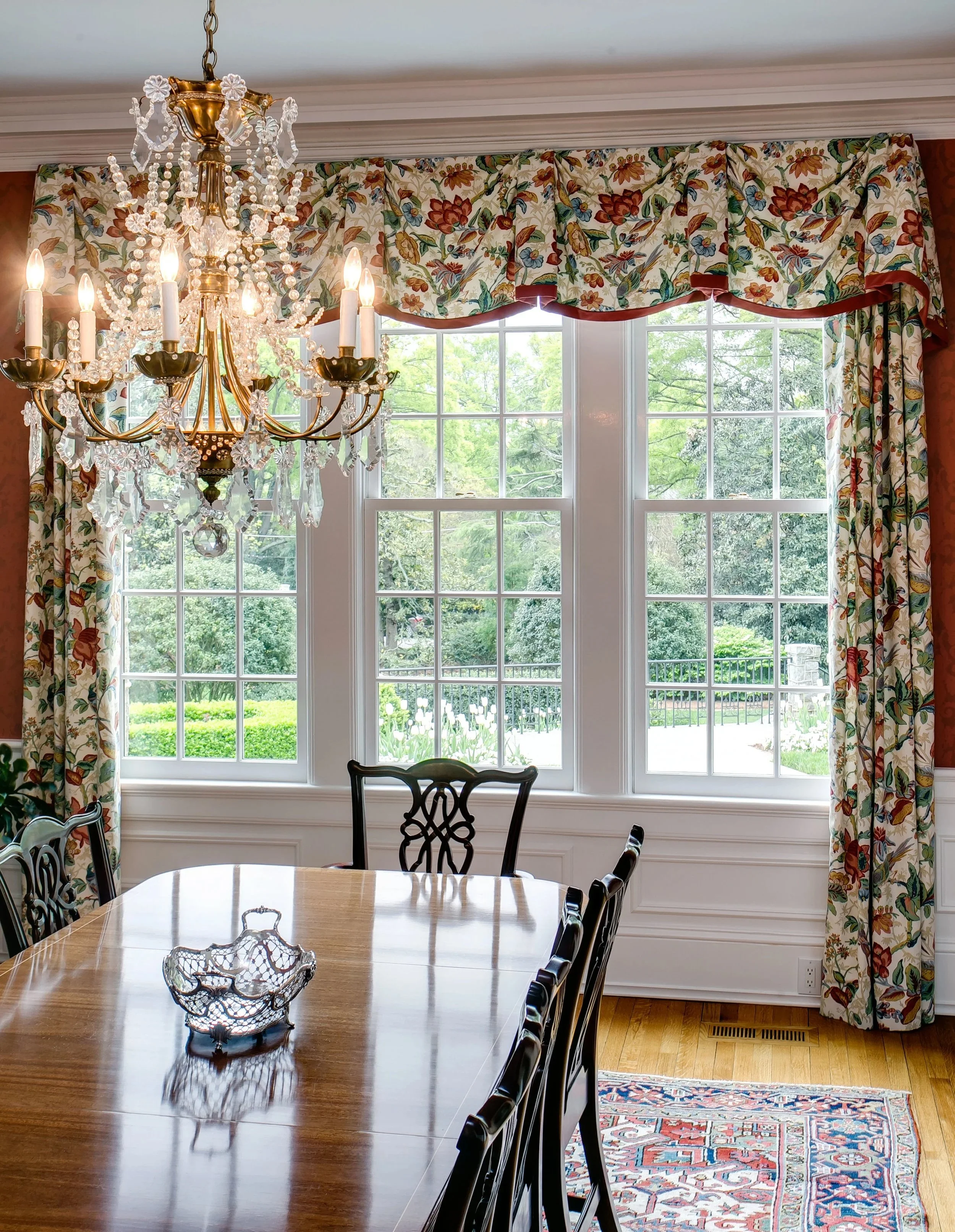 A dining room with a wooden table, decorative chairs, a chandelier, large window with floral curtains, and a view of greenery outside.