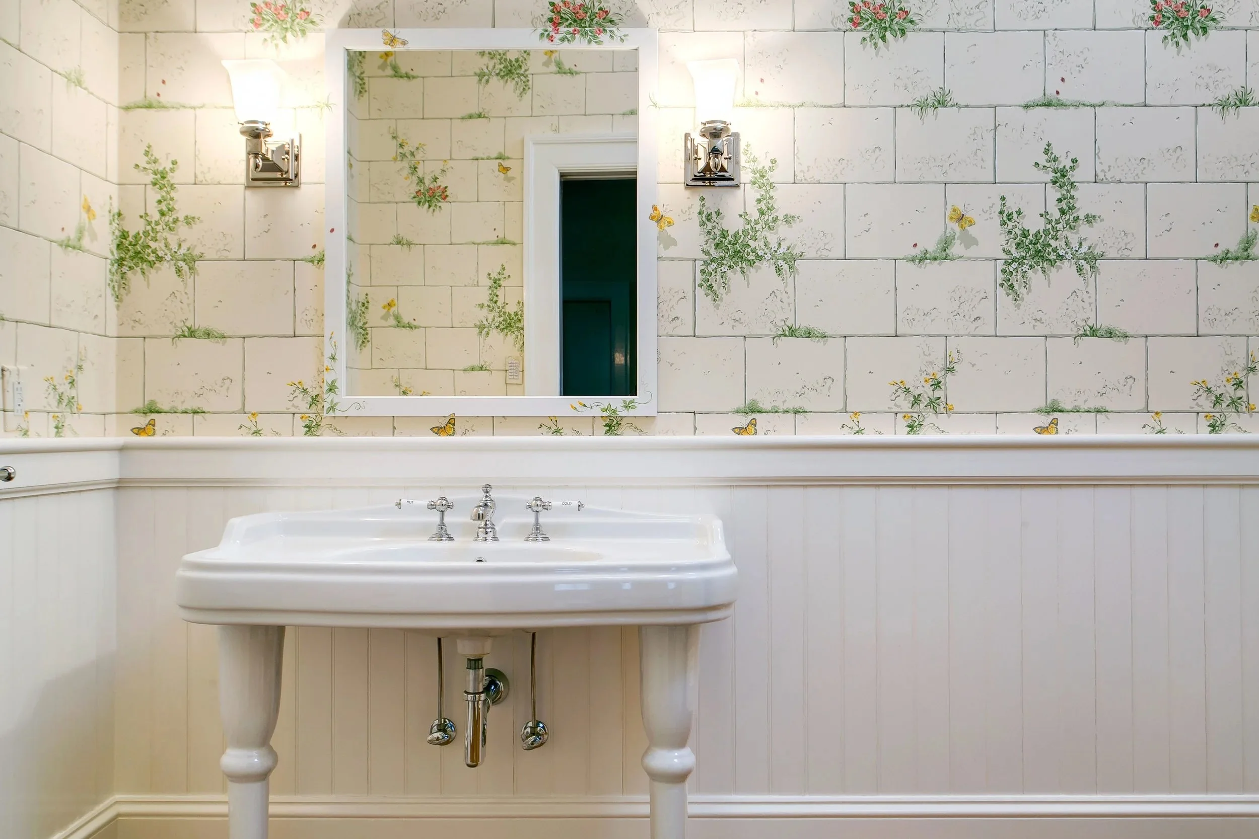 Bathroom with a white sink, a mirror with a white frame, wall-mounted light fixtures, and wallpaper with a floral and butterfly pattern.