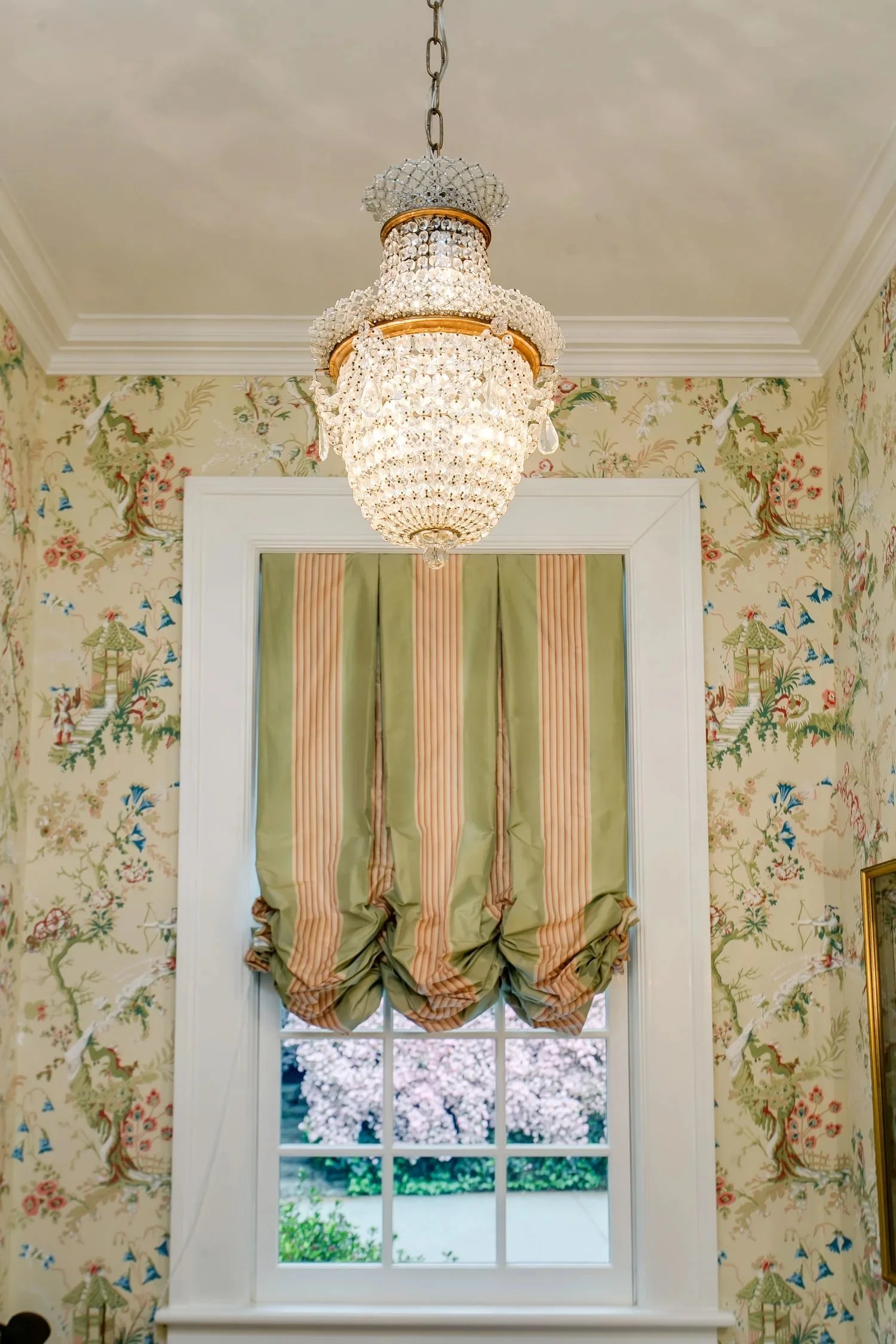A window with a green and red striped curtain, and a view of a flowering tree and greenery outside. The window is framed by wallpaper with a floral and landscape motif. A crystal chandelier hangs from the ceiling above the window.