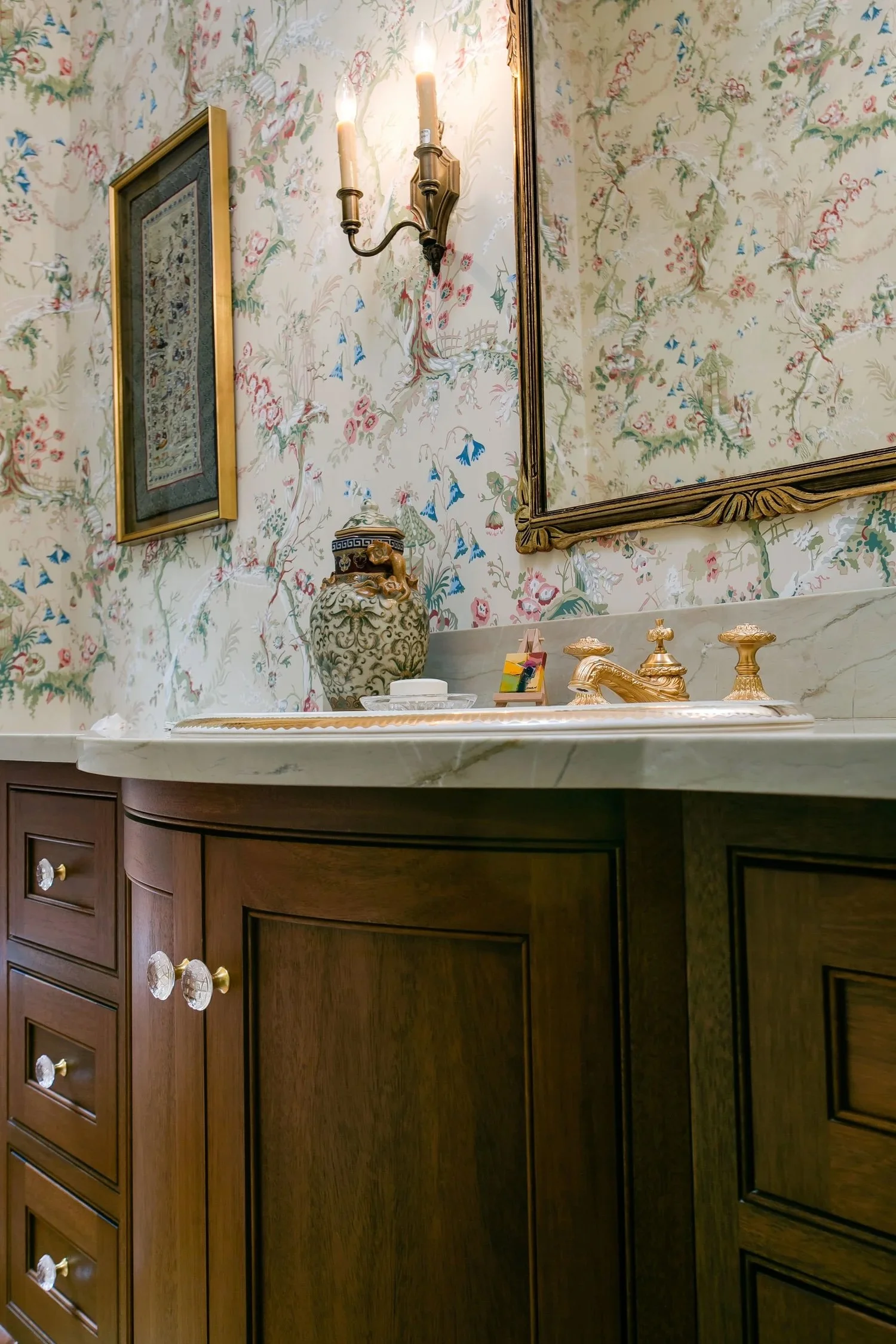 A bathroom vanity with a marble countertop featuring a gold faucet, decorative gold containers, and a leopard-print jar. The background has wallpaper with a floral and nature scene, framed pictures, and a brass wall sconce with candles.