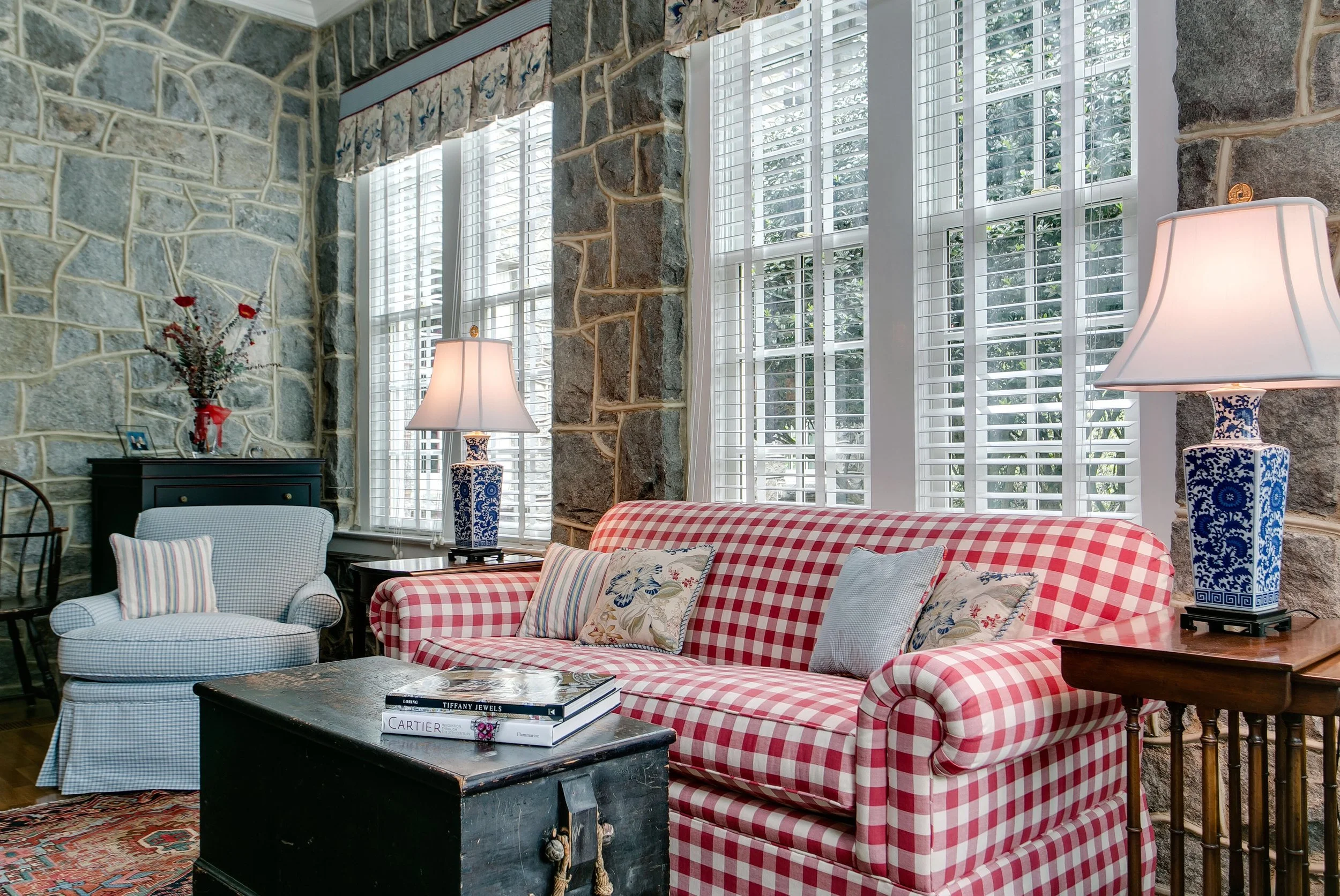 Living room with stone walls, large windows with white blinds, and furniture including a checkered red and white sofa, a white armchair, and a dark coffee table with books. There are table lamps with blue and white bases and floral cushions on the so