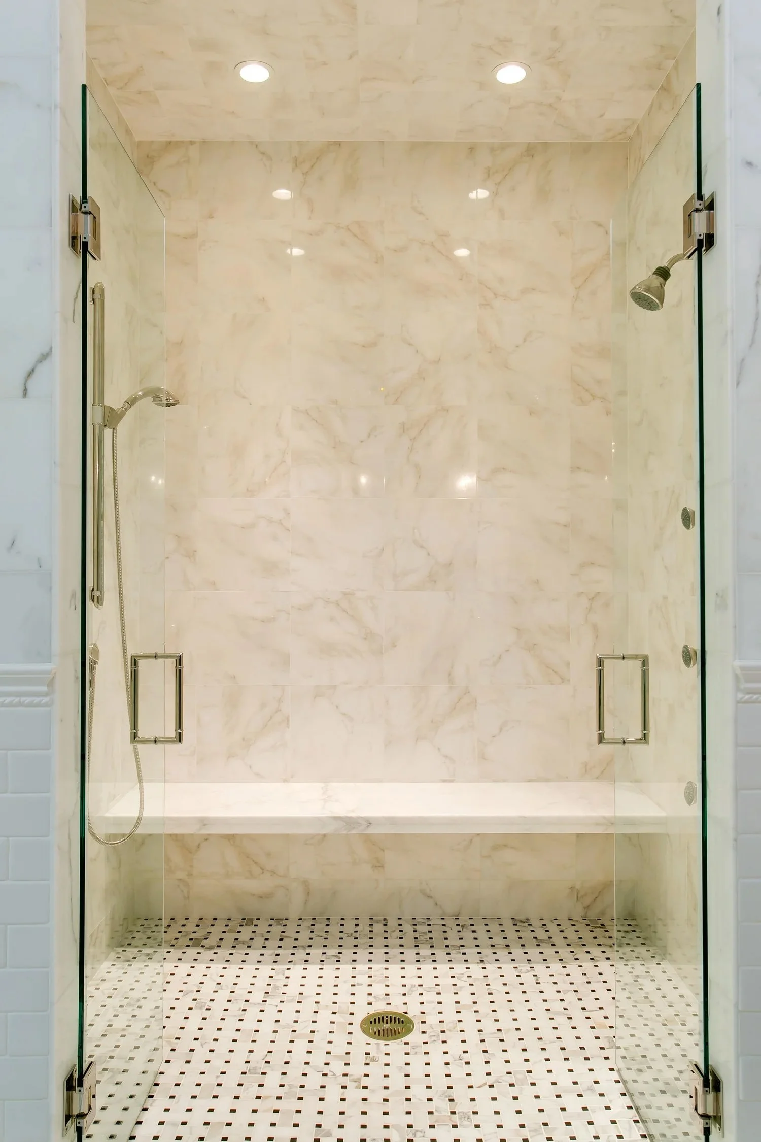 A modern walk-in shower with beige marble walls, a glass enclosure with metal handles, ceiling lights, two showerheads, a handheld shower, and a tiled floor with a drain in the center.