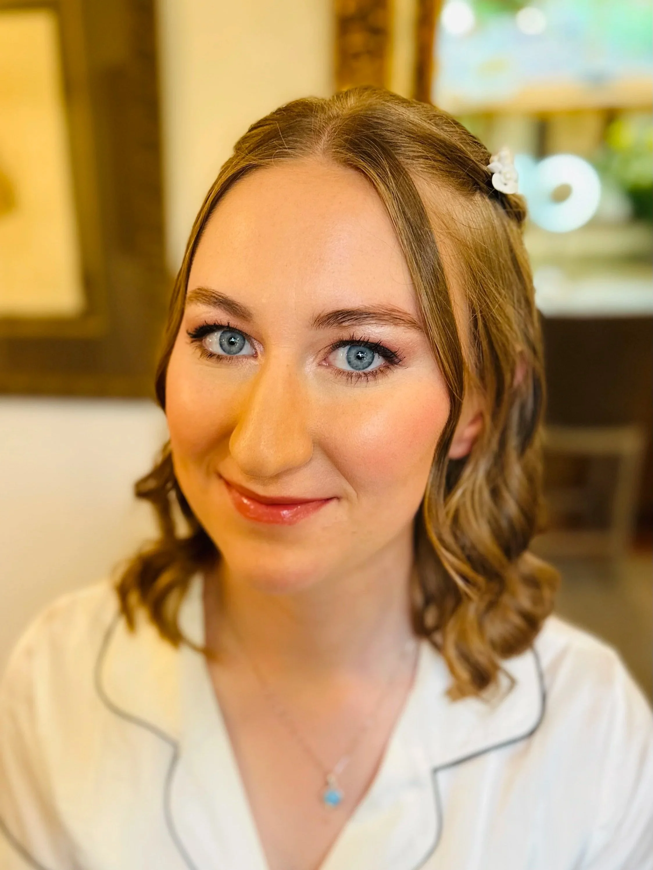 Close-up of a woman with light brown hair and blue eyes, wearing a white top with black piping and a blue pendant necklace, smiling softly.