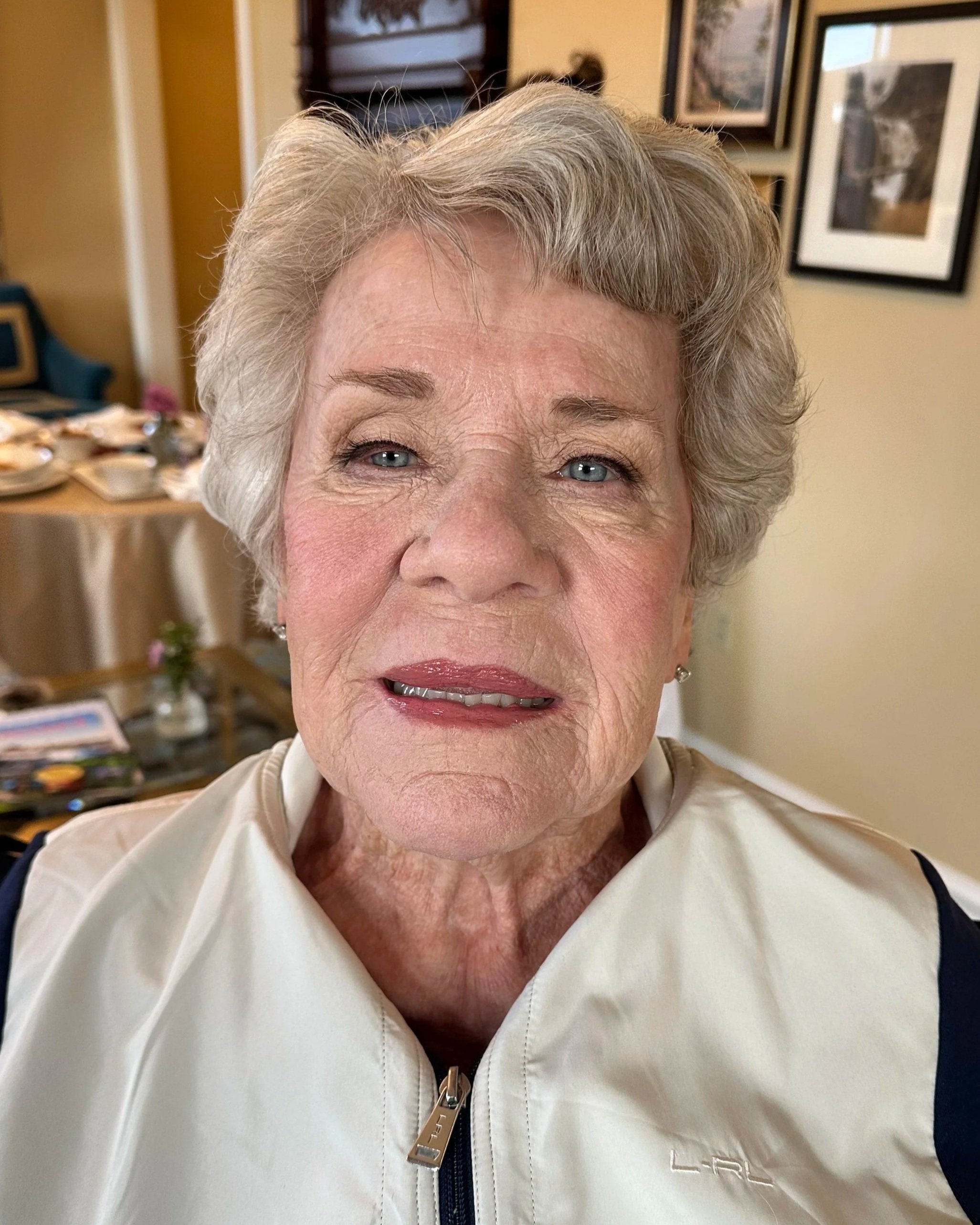 Close-up of an elderly woman with short gray hair and blue eyes, smiling slightly, wearing a white jacket with a zipper. In the background, there are framed pictures on the wall and a table with plates and glasses.