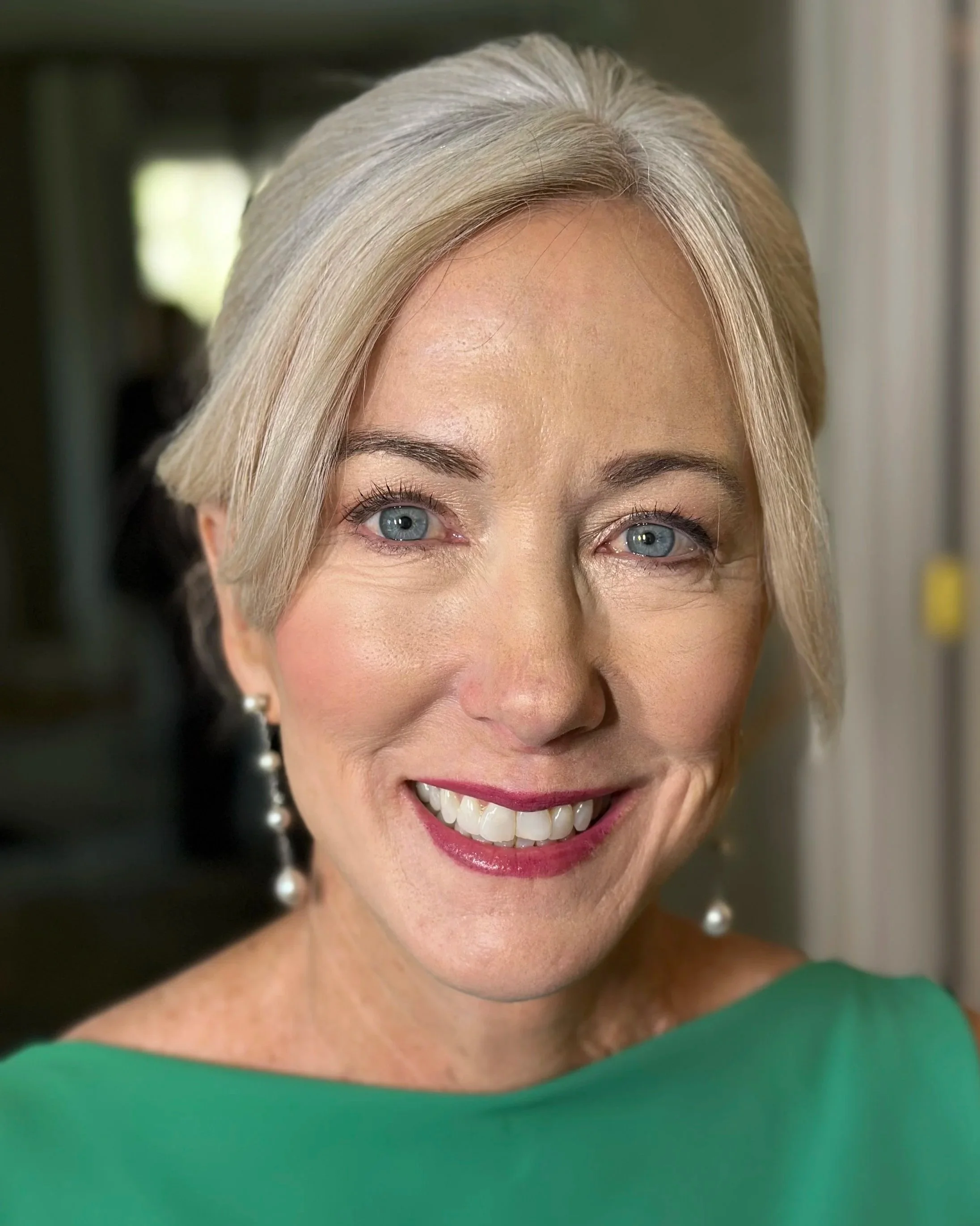 Close-up of a woman with blonde hair, blue eyes, wearing pearl earrings and a green dress, smiling.