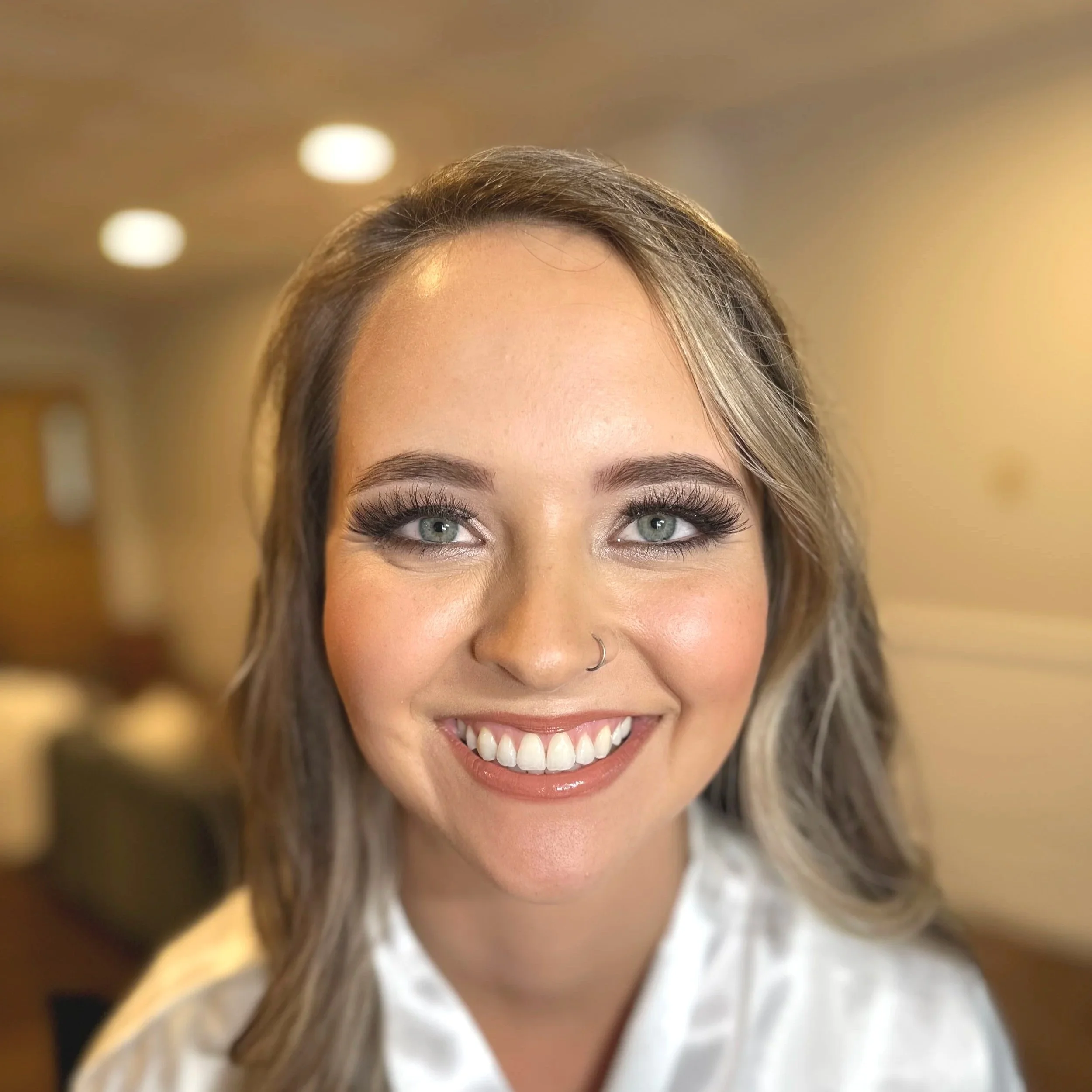 Close-up of a woman with blue eyes, long eyelashes, smiling, with light brown hair and a nose piercing.