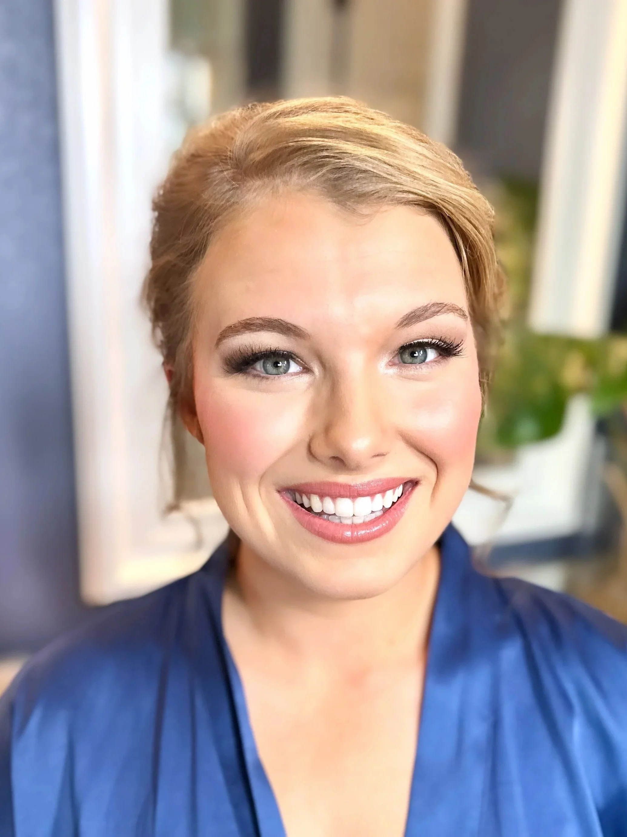 Close-up of a smiling woman with blonde hair, blue eyes, wearing blue clothing, in an indoor setting.
