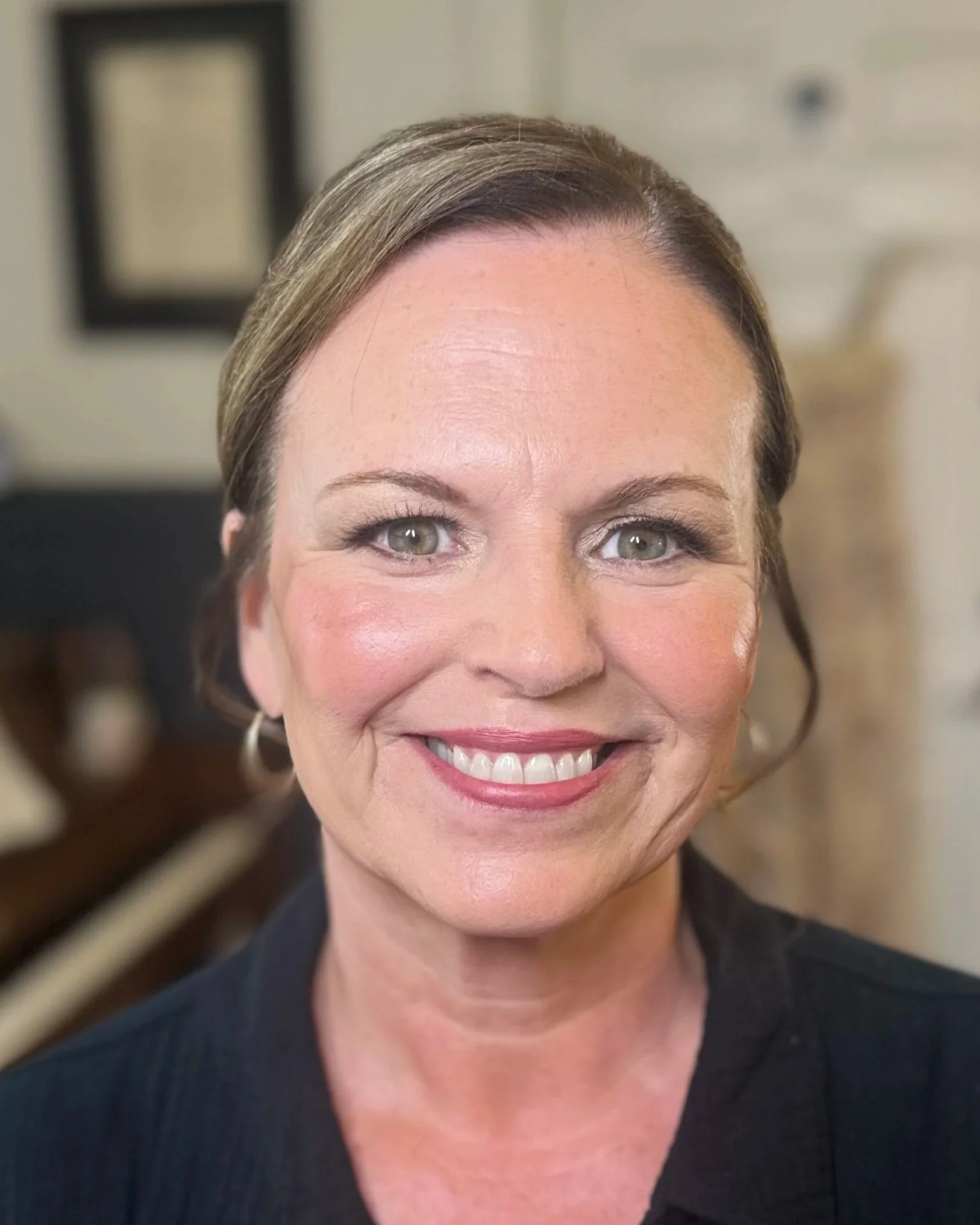 A smiling woman with short brown hair and green eyes, wearing a black top and earrings, is in a room with framed pictures on the wall behind her.