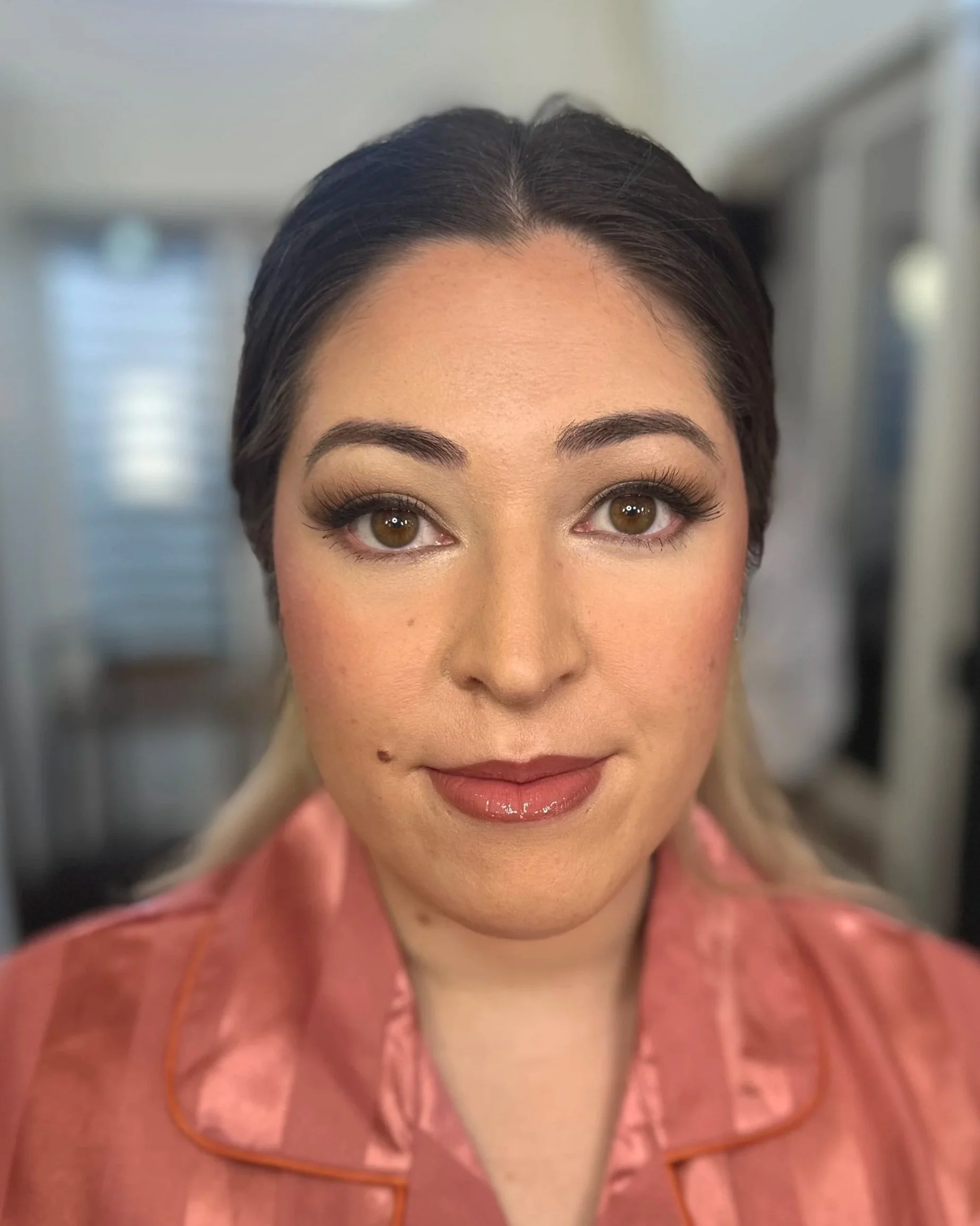 Close-up of a woman's face showing her brown eyes, well-groomed eyebrows, and dark hair styled back. She is wearing a pink shirt and has makeup on, including eyeliner, mascara, and lipstick.