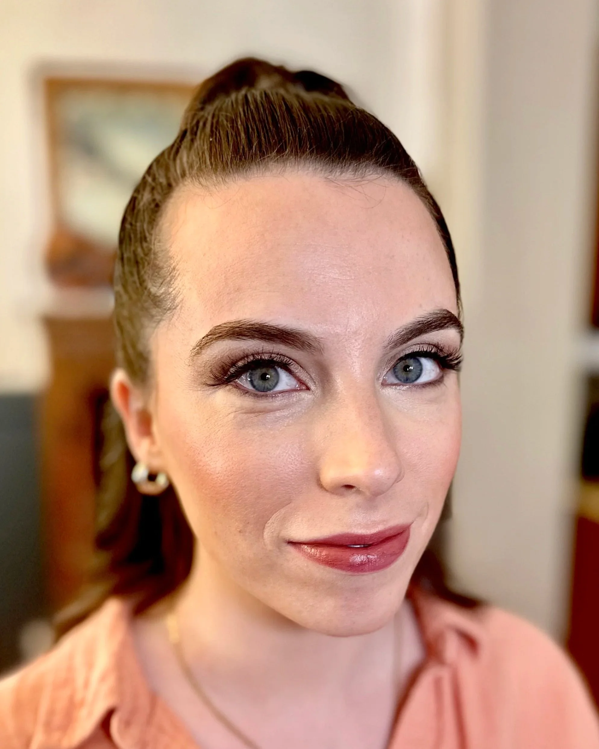 Close-up of a smiling woman with light blue eyes, brown hair styled in an elegant updo, wearing makeup with defined eyebrows and pink lipstick, wearing a peach-colored top inside a room.