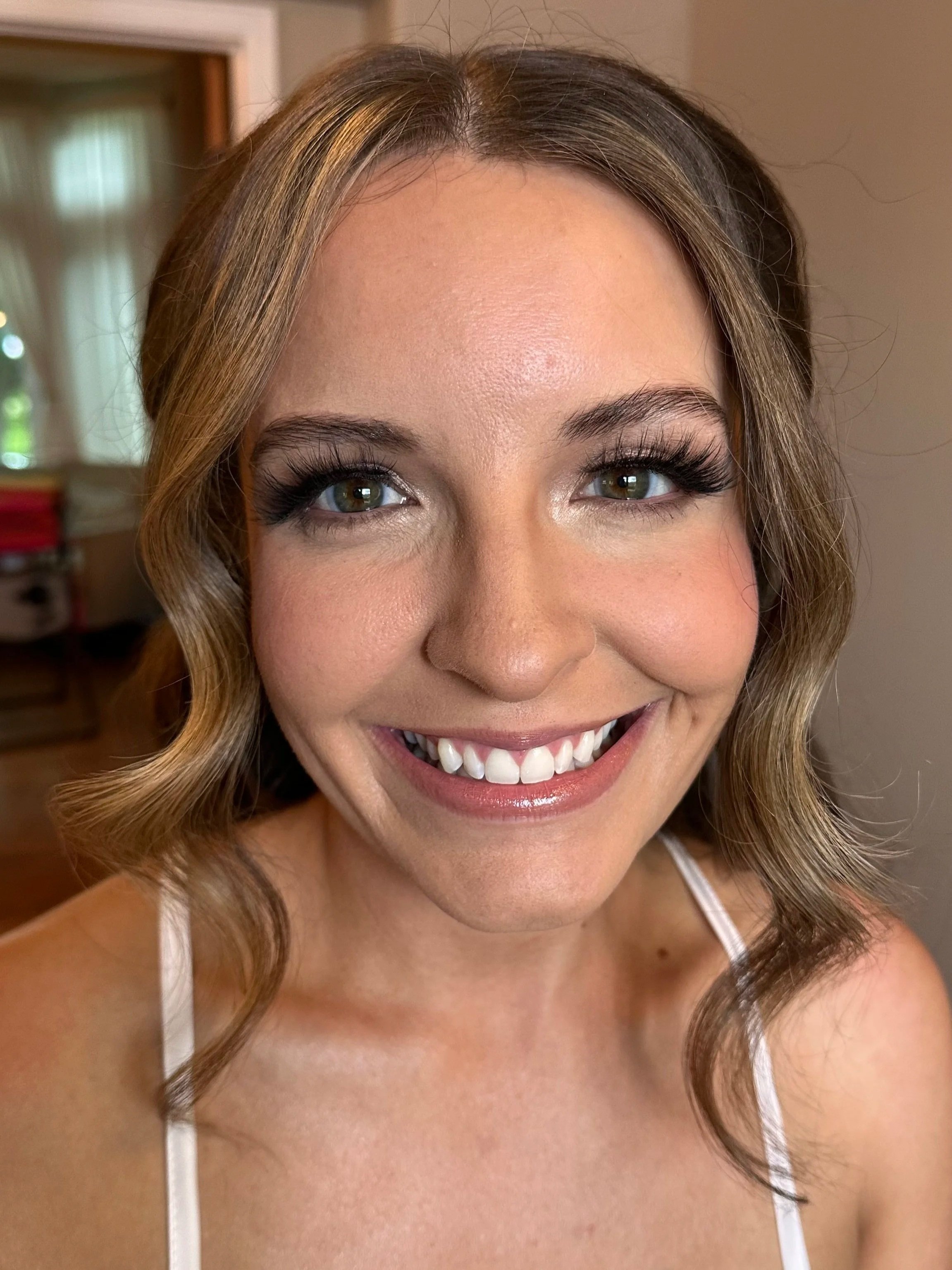A smiling woman with brown hair, blue eyes, and makeup, taking a selfie in a room with a blurred background.