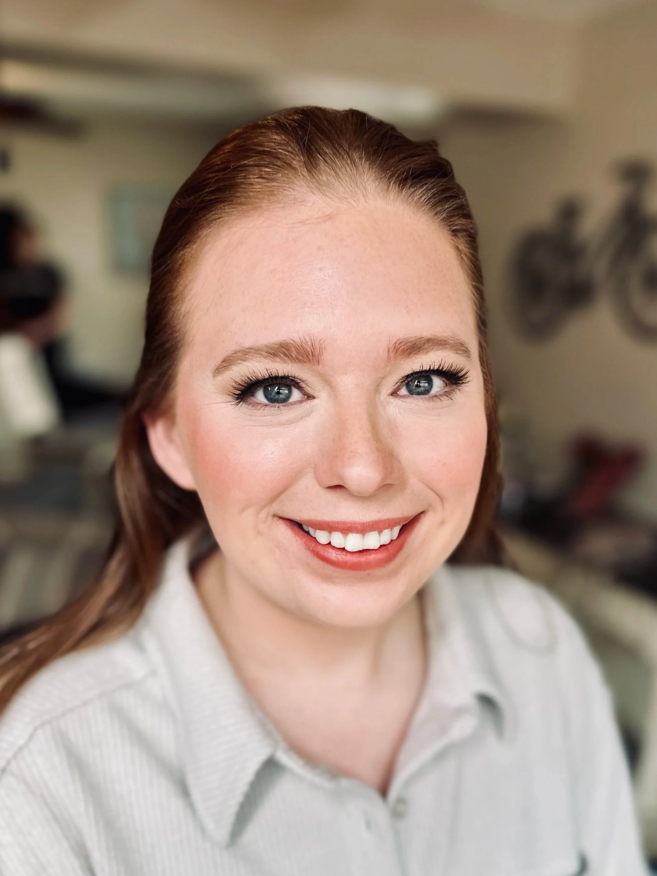 Close-up of a smiling woman with blue eyes, red hair, and light skin wearing a light-colored sweater.