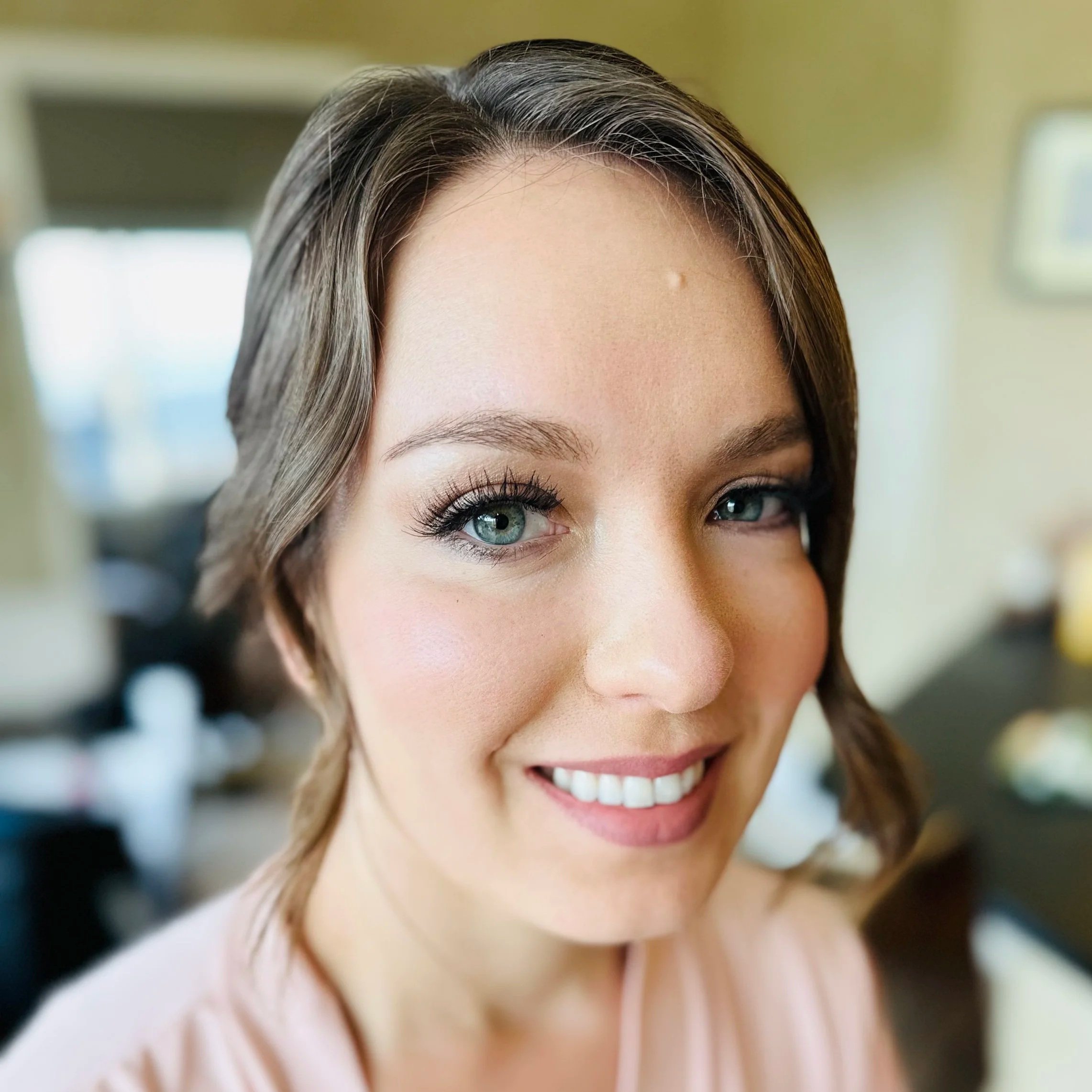 Close-up of a smiling woman with light brown hair, blue eyes, and makeup in an indoor setting with blurred background.