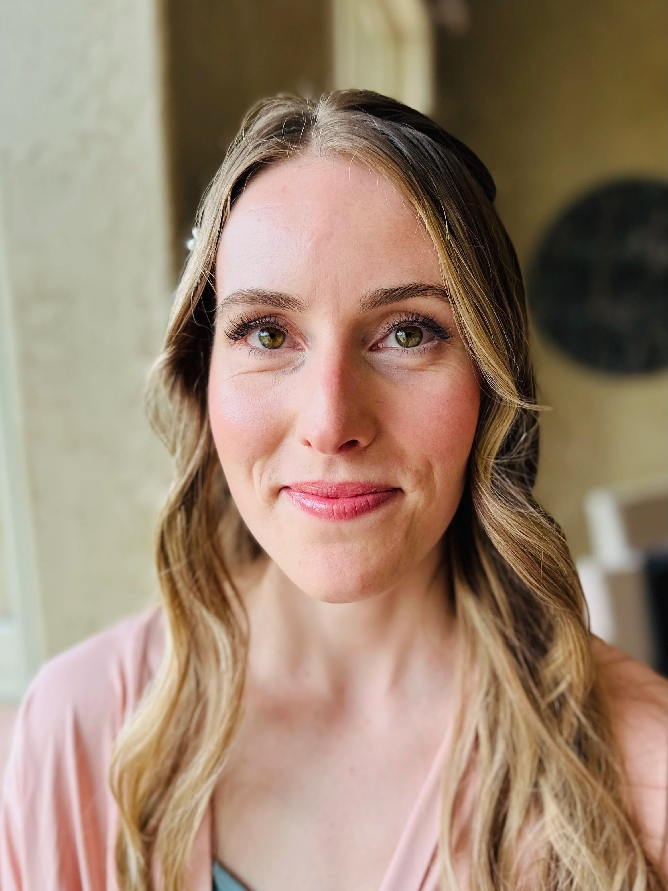 A close-up of a woman with long, wavy blonde hair and light skin, smiling softly with hazel eyes, wearing a pink blouse.
