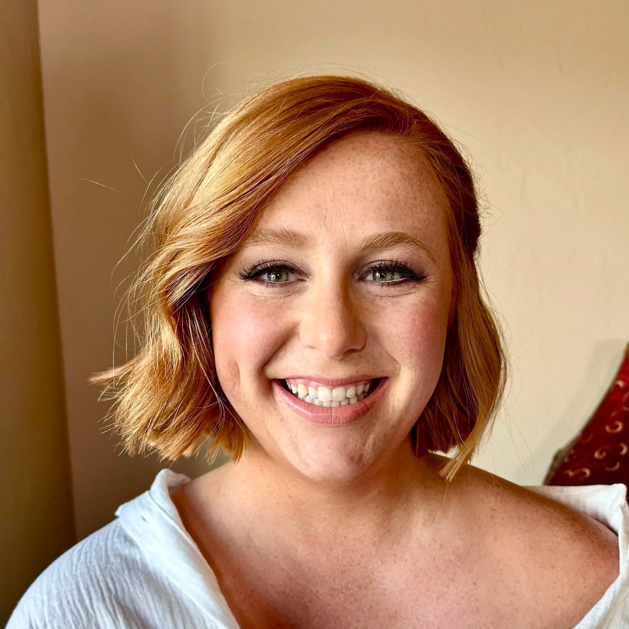 Close-up of a smiling woman with red hair, freckles, and green eyes, wearing a white top.