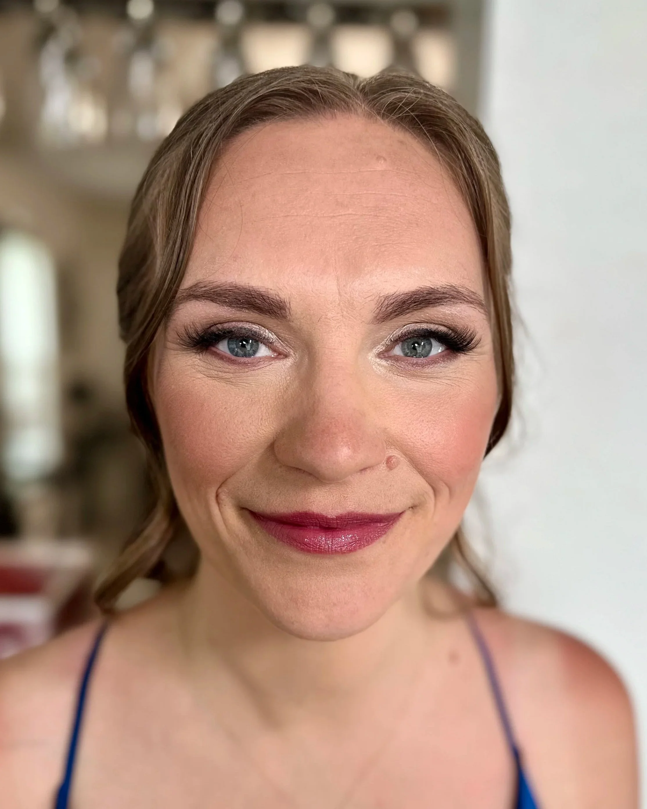 Close-up of a woman smiling, showing her eyes and part of her face, with light makeup, wearing a dark strap top, with brown hair styled in loose waves.