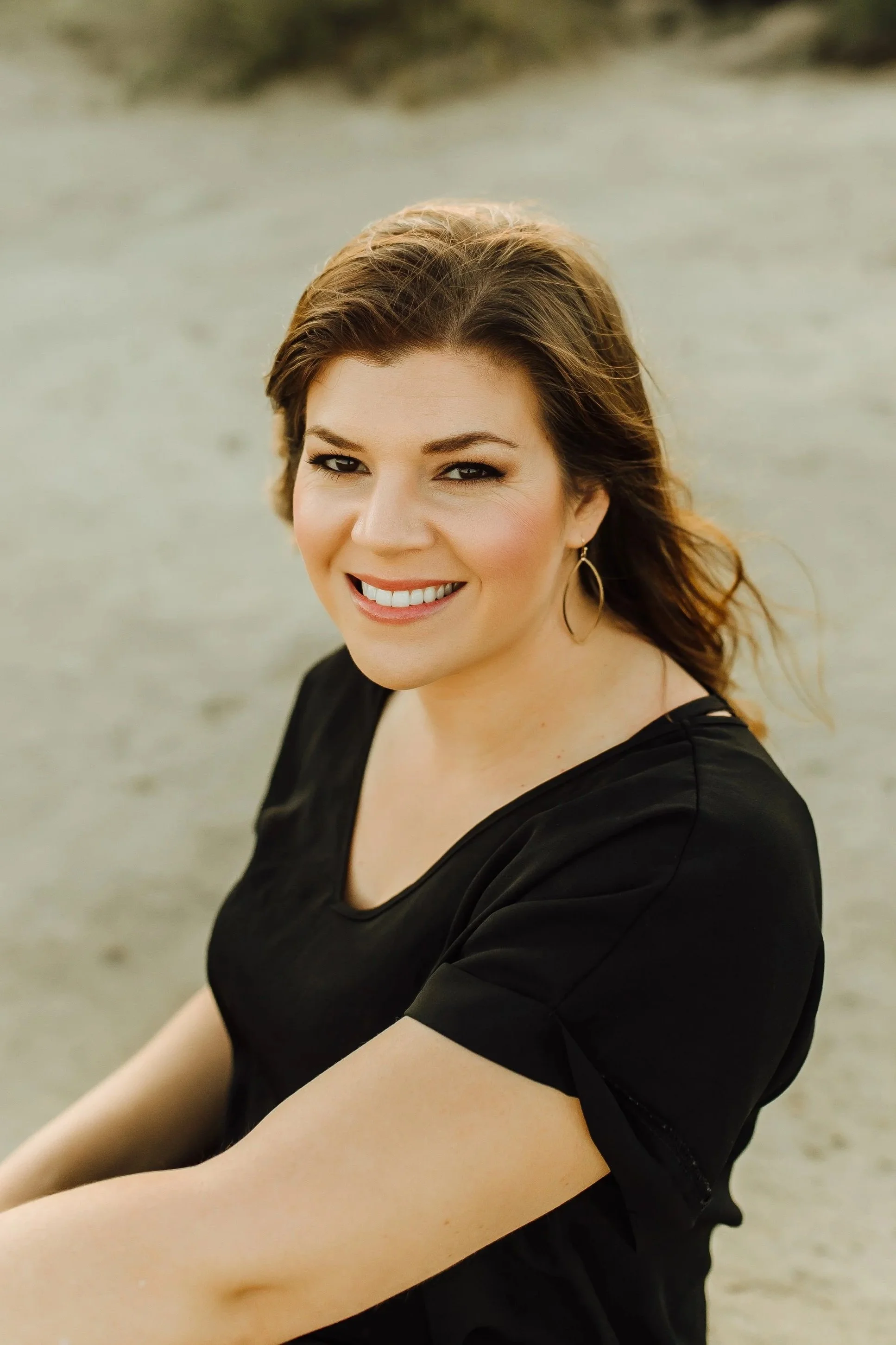 Close-up of a smiling woman with light skin, brown hair, and hoop earrings, wearing a black top, sitting outdoors on a sandy surface.