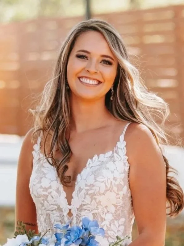 Smiling woman in a white lace dress holding a bouquet of blue flowers, outdoors with a wooden fence in the background.