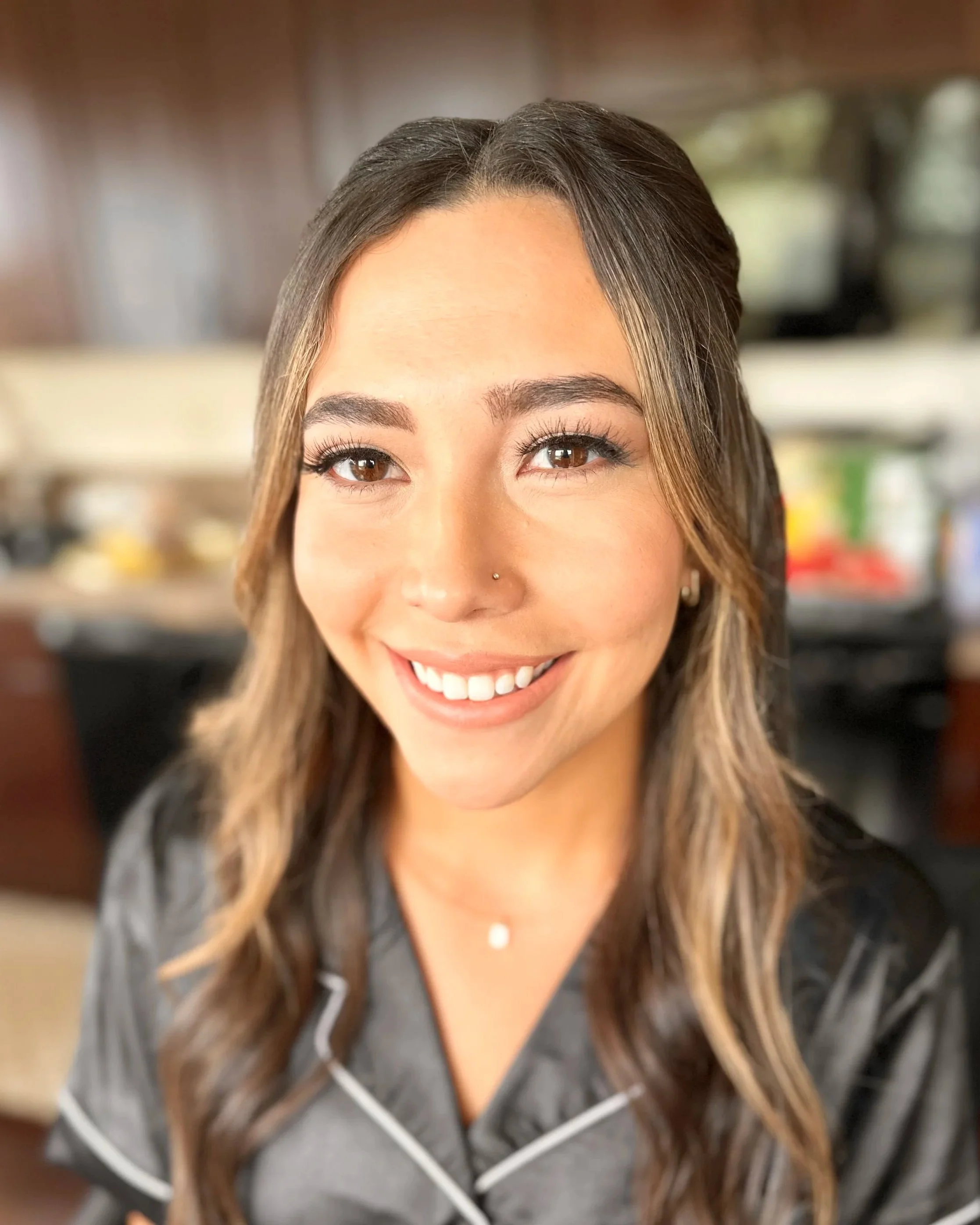 Close-up of a smiling woman with long wavy brown hair, wearing a gray top with white piping, and a delicate necklace, in an indoor setting.