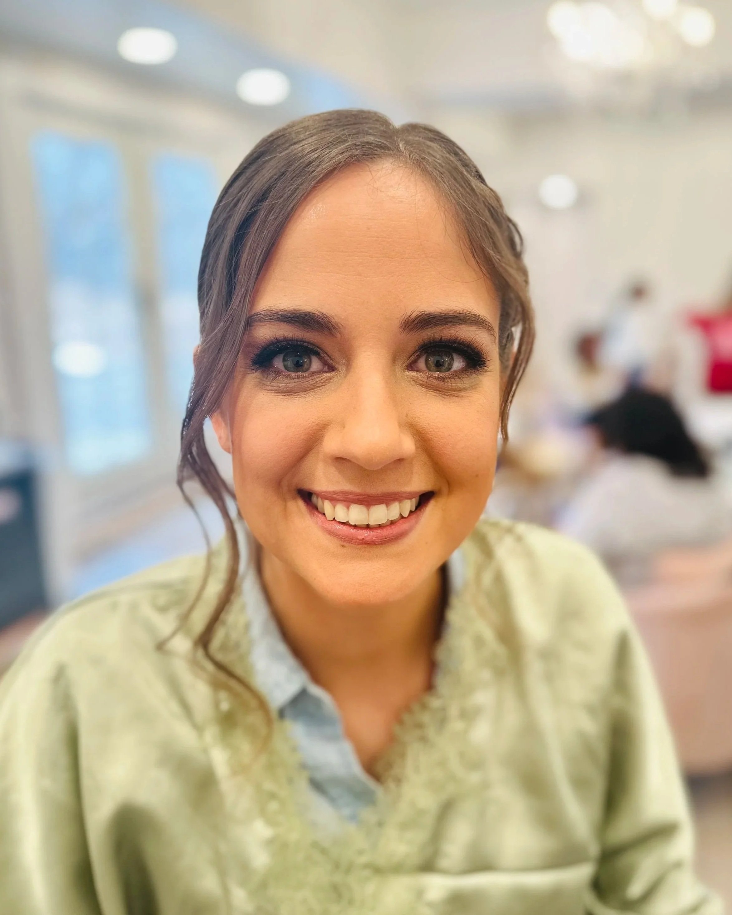 Close-up of a woman smiling, with visible upper face, brown hair, wearing a light yellow sweater, indoors with blurred background.