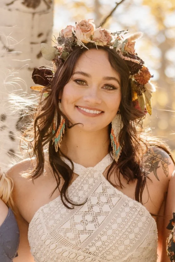 A woman smiling outdoors wearing a flower crown, intricate lace dress, and colorful feather earrings.