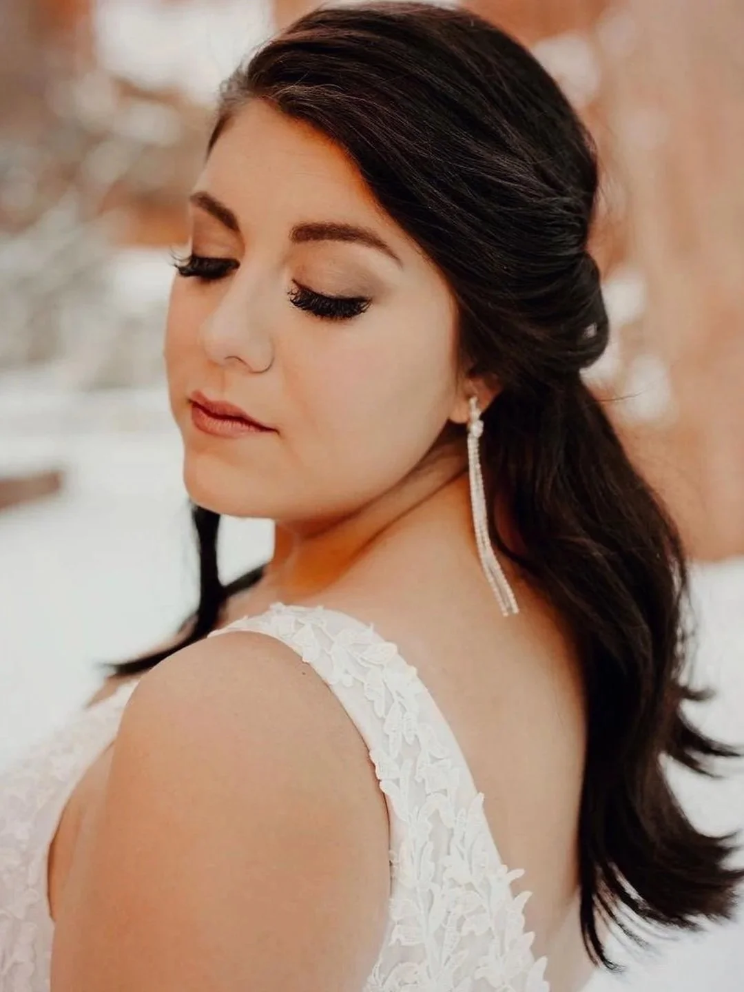 Close-up of a woman with dark, wavy hair styled to one side, wearing long, dangling earrings, and a white lace dress, with her eyes closed and a serene expression.