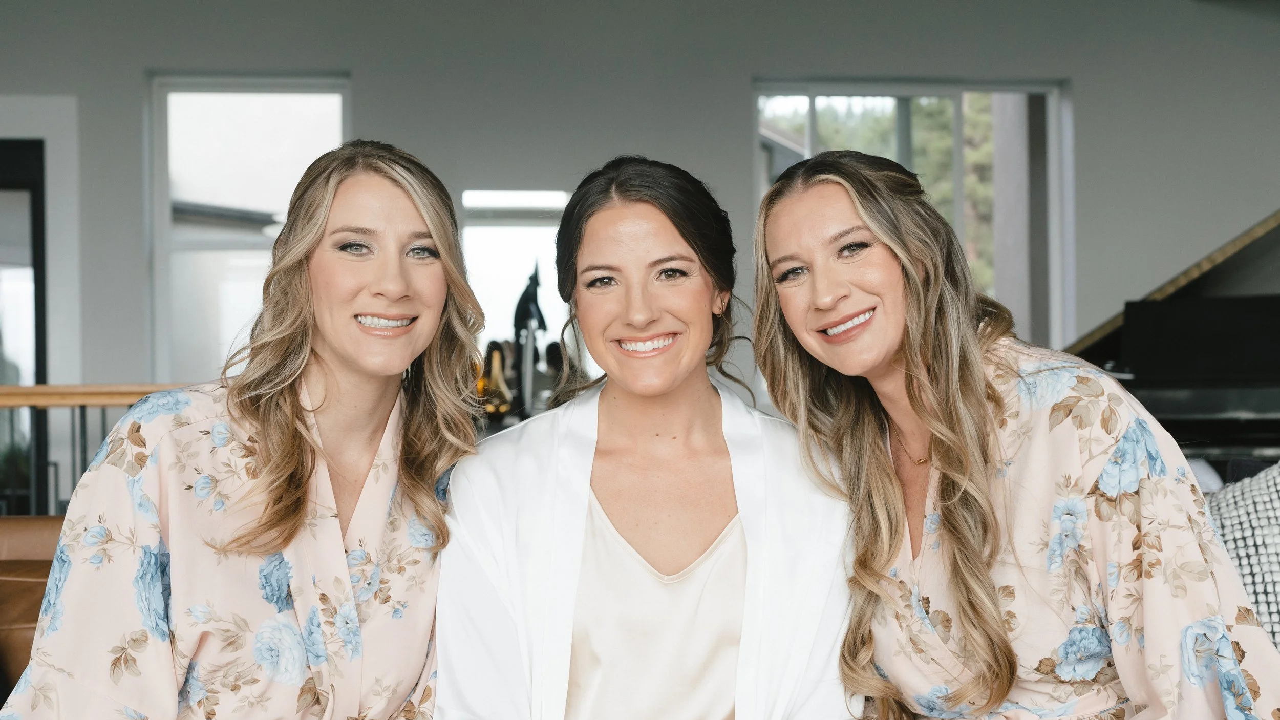 Three women smiling indoors, one in the middle with dark hair, and the other two with blonde hair on either side, wearing floral robes.