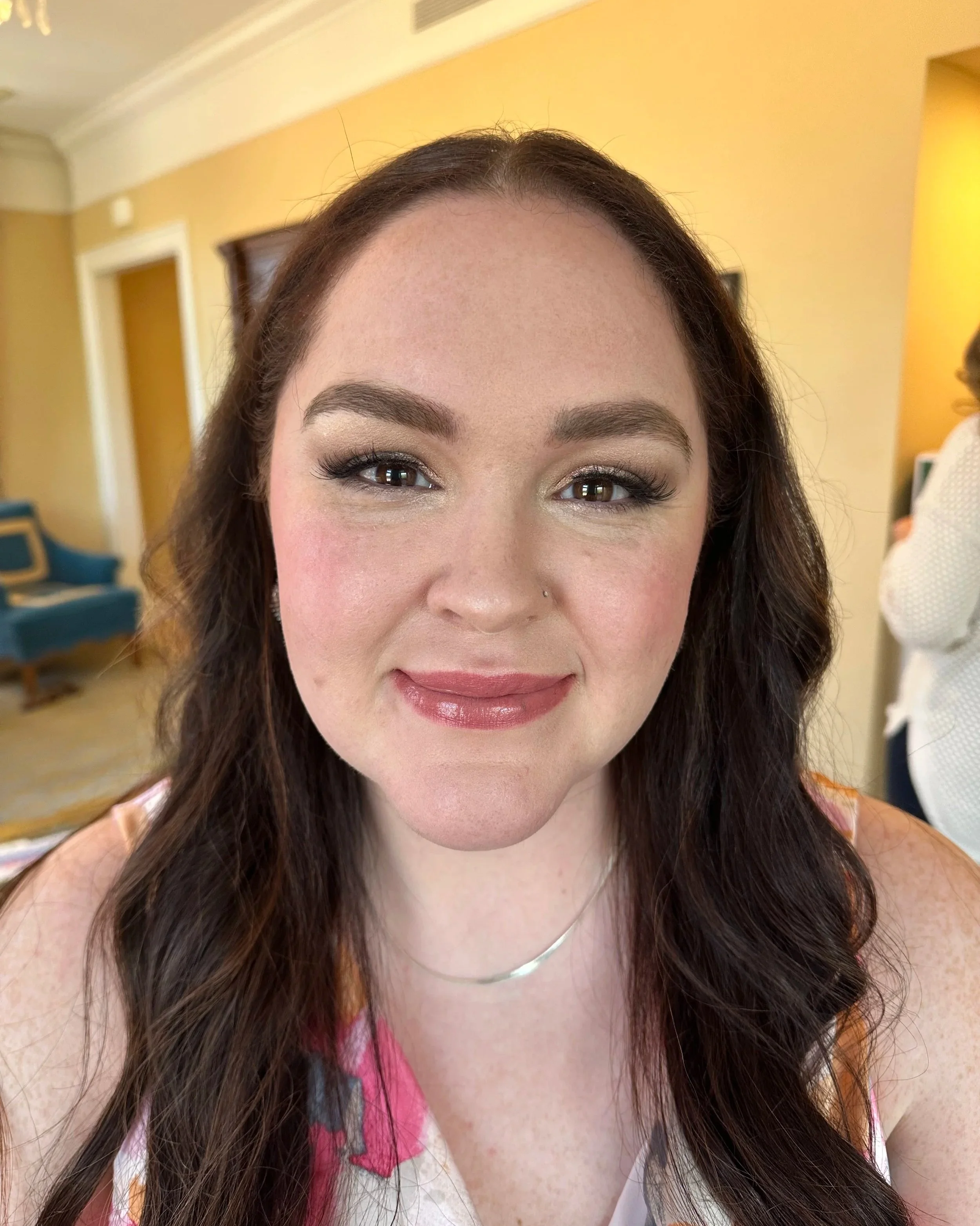 Close-up of a smiling woman with long wavy brown hair, wearing makeup including eyeshadow, mascara, and lipstick, in a warmly lit room with yellow walls and furniture in the background.