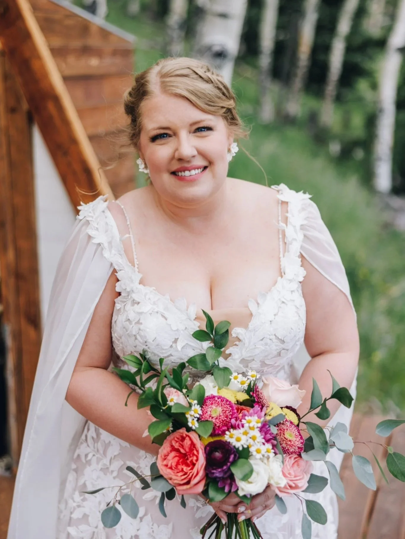 A smiling woman dressed in a white lace wedding gown holding a colorful bouquet of flowers outdoors, with a wooden staircase and trees in the background.