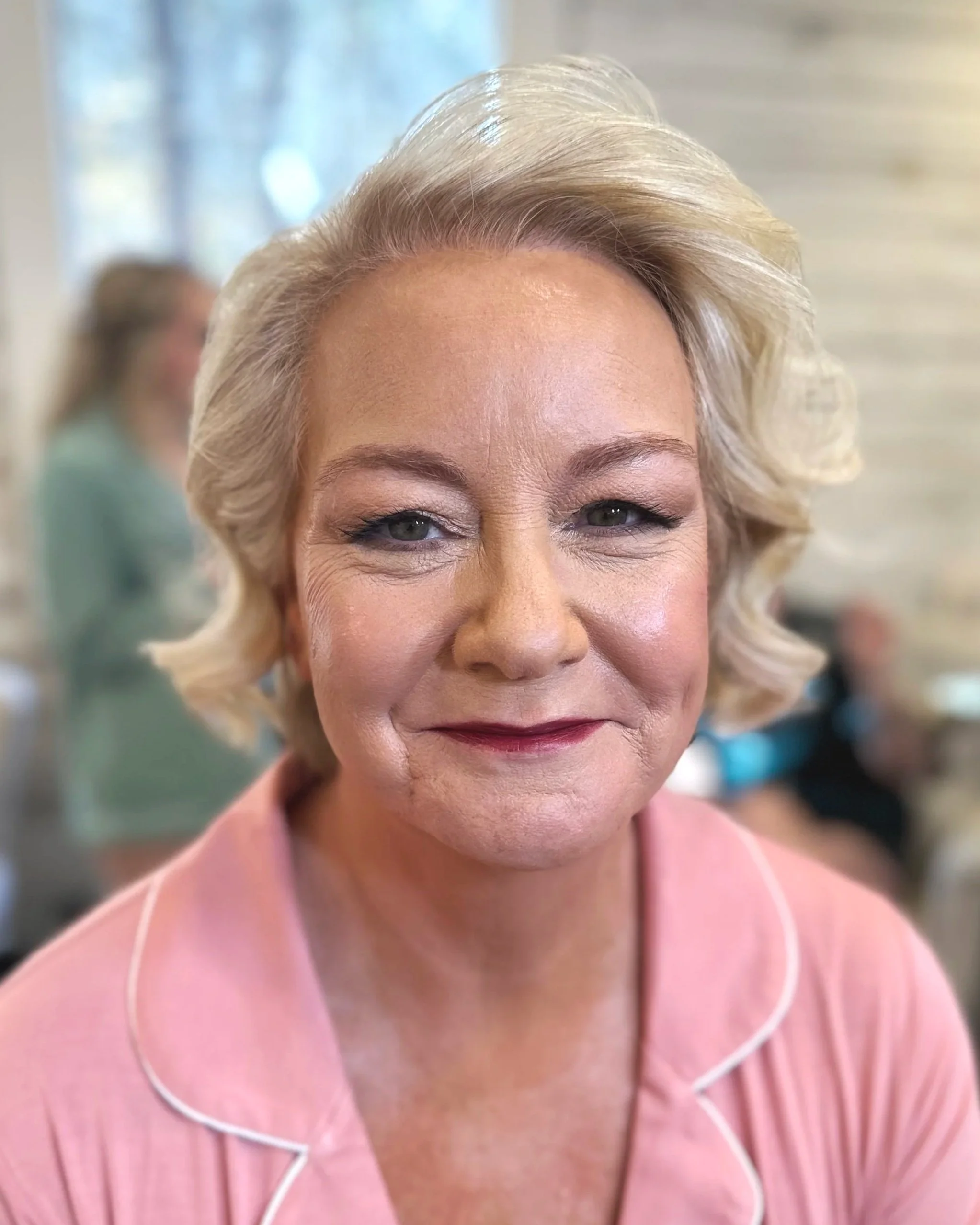 Close-up of a woman with blonde, styled hair, wearing light pink pajamas with white piping, smiling softly.