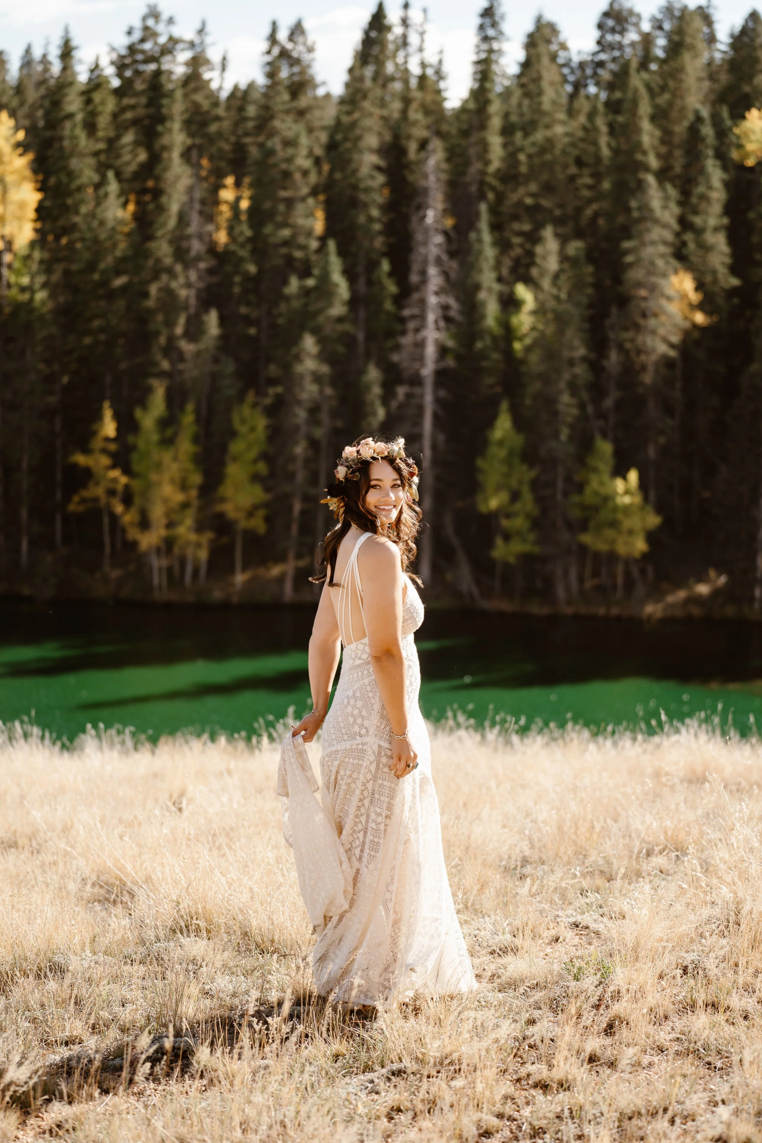 A woman in a white lace dress with a floral crown standing in a field of tall, dry grass near a lake, with a dense forest of tall pine trees in the background.