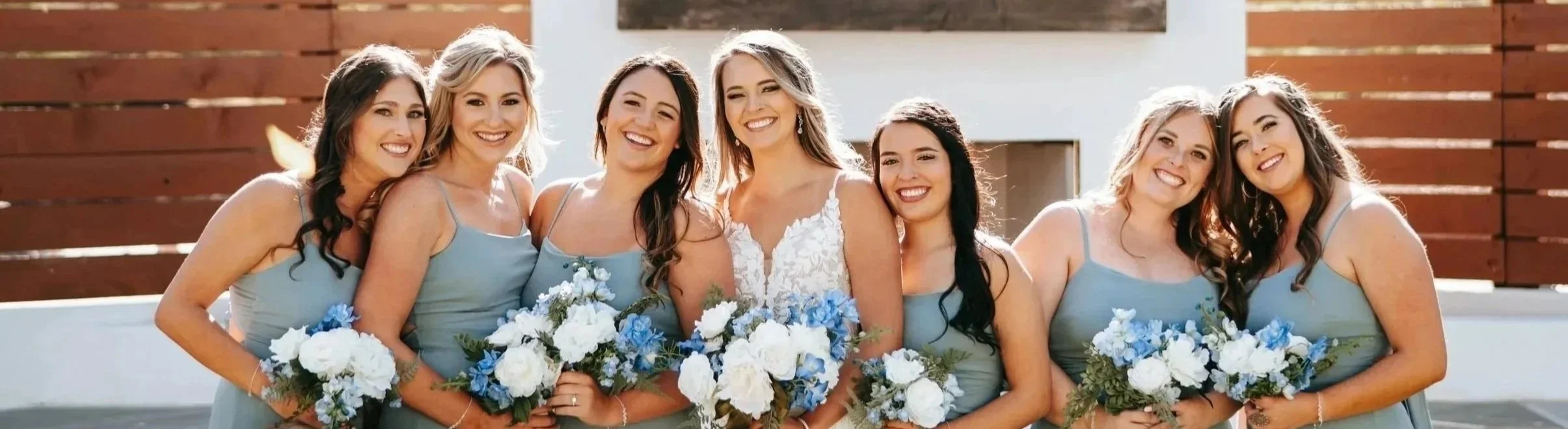 Group of seven women smiling, wearing bridesmaid dresses, holding bouquets of white and blue flowers, outdoors with wooden fence in background.