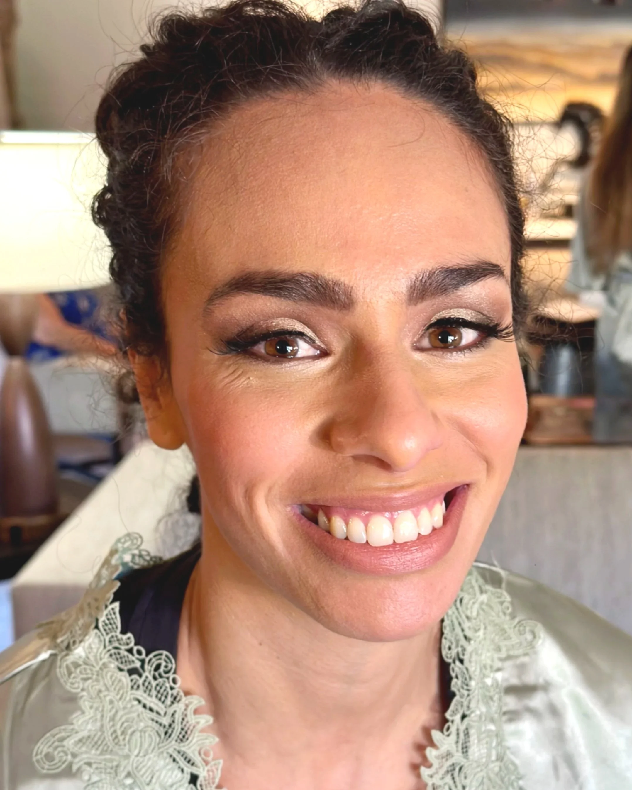 Close-up of a smiling woman with dark curly hair, wearing a light-colored lace top, in a well-lit indoor setting.