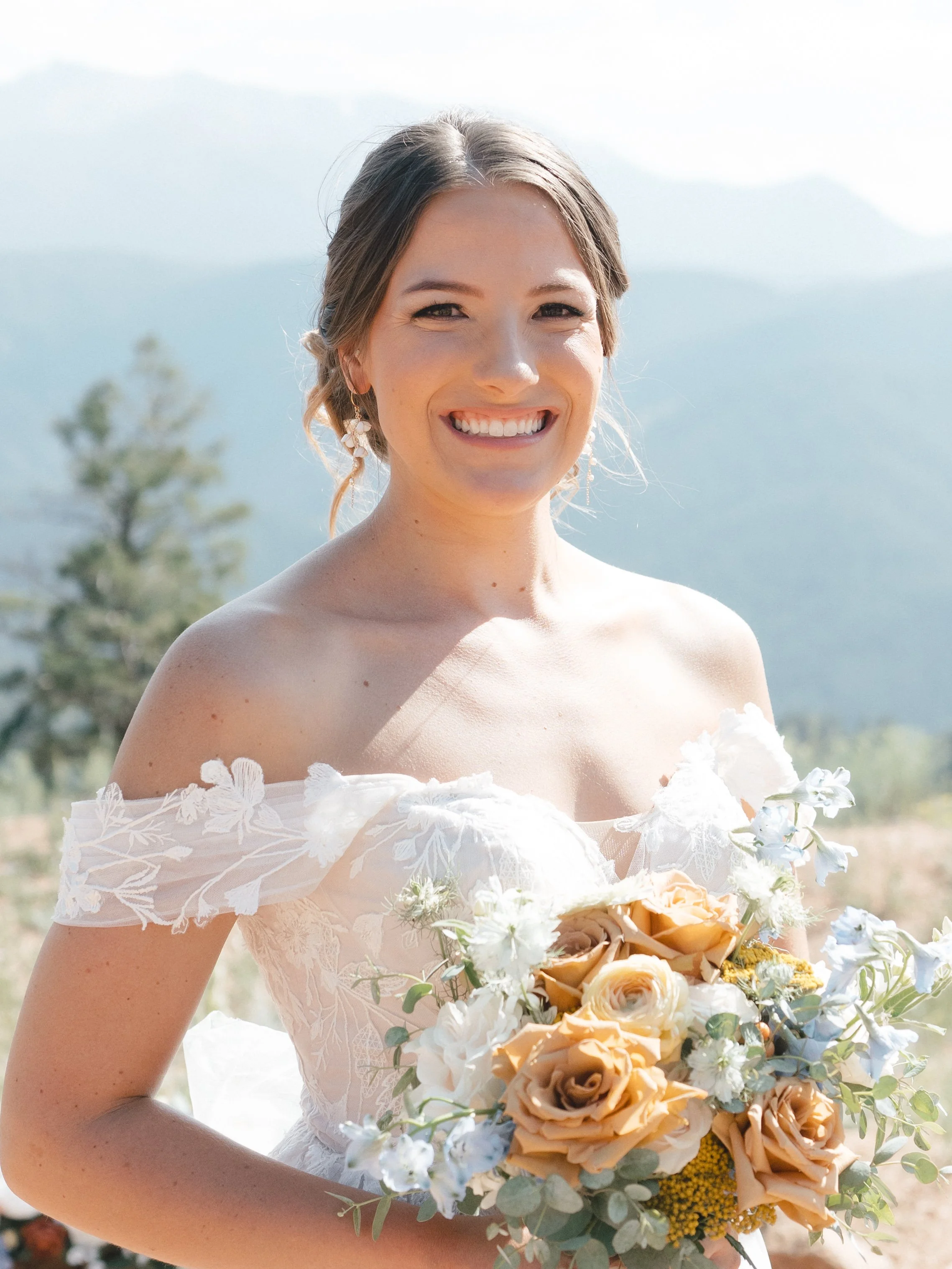 A smiling woman in a wedding dress holding a large bouquet of flowers outdoors with mountains and trees in the background.