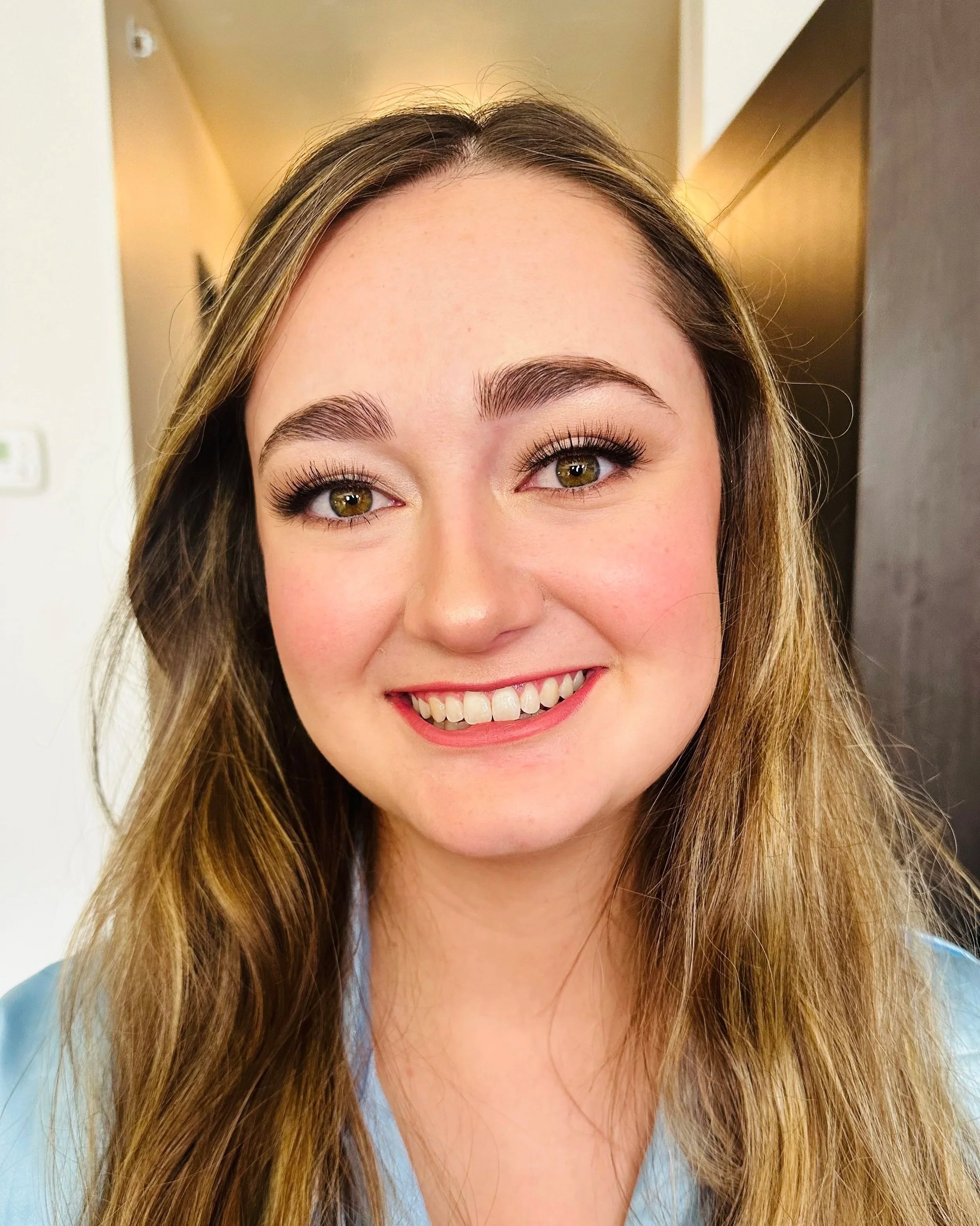 Close-up of a smiling woman's face showing her eyes, eyebrows, and part of her nose and mouth. She has long, wavy brown hair and is wearing light makeup.