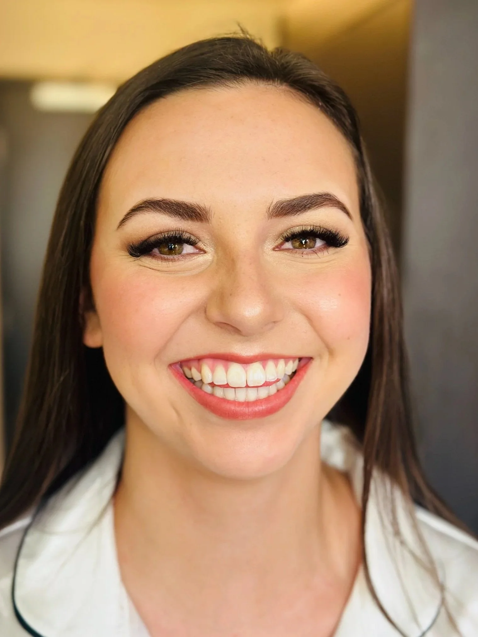 Close-up of a smiling woman with long brown hair, wearing makeup and a white top with black piping.