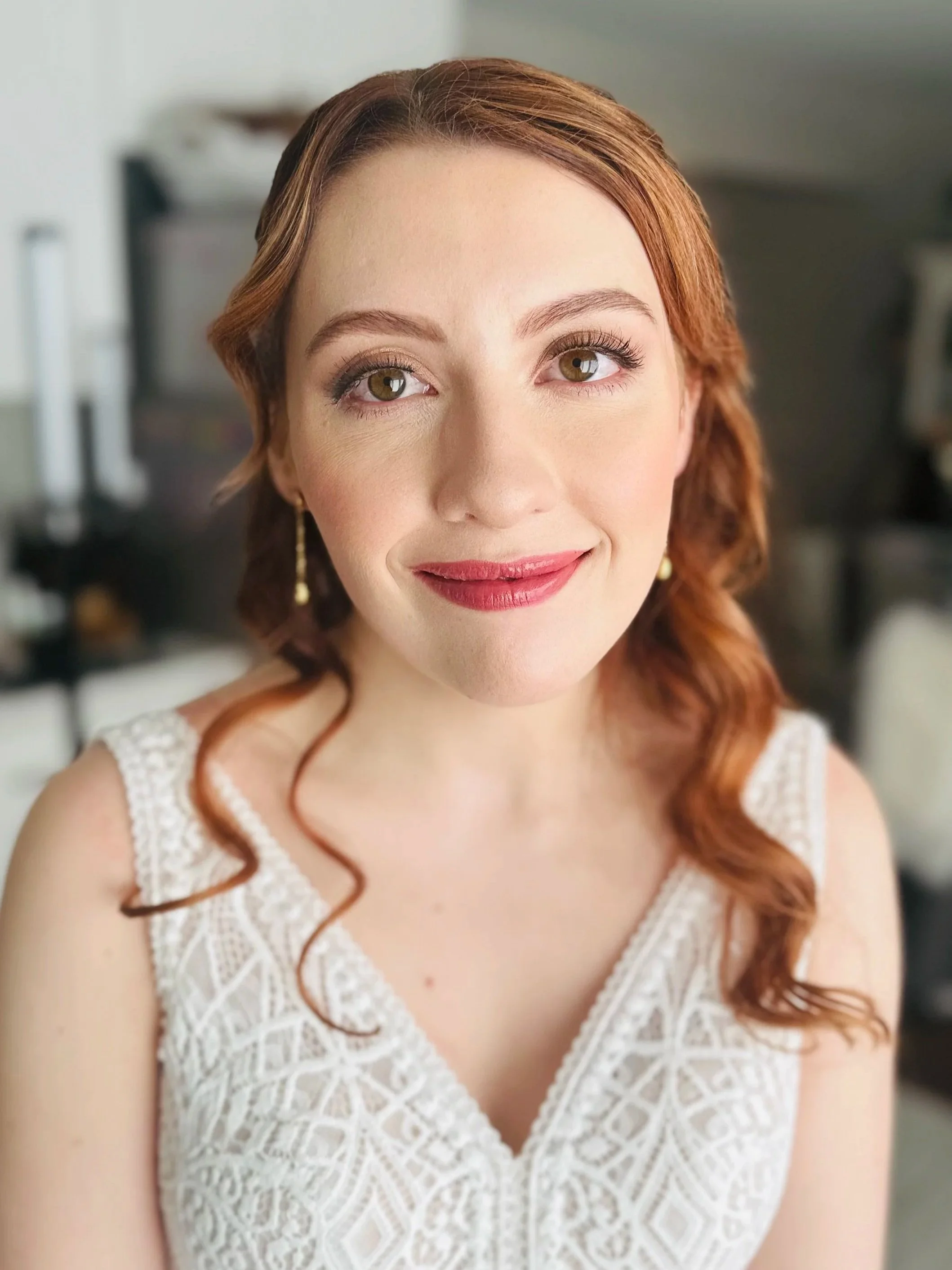A woman with red hair, hazel eyes, and fair skin is smiling while wearing a white lace dress.