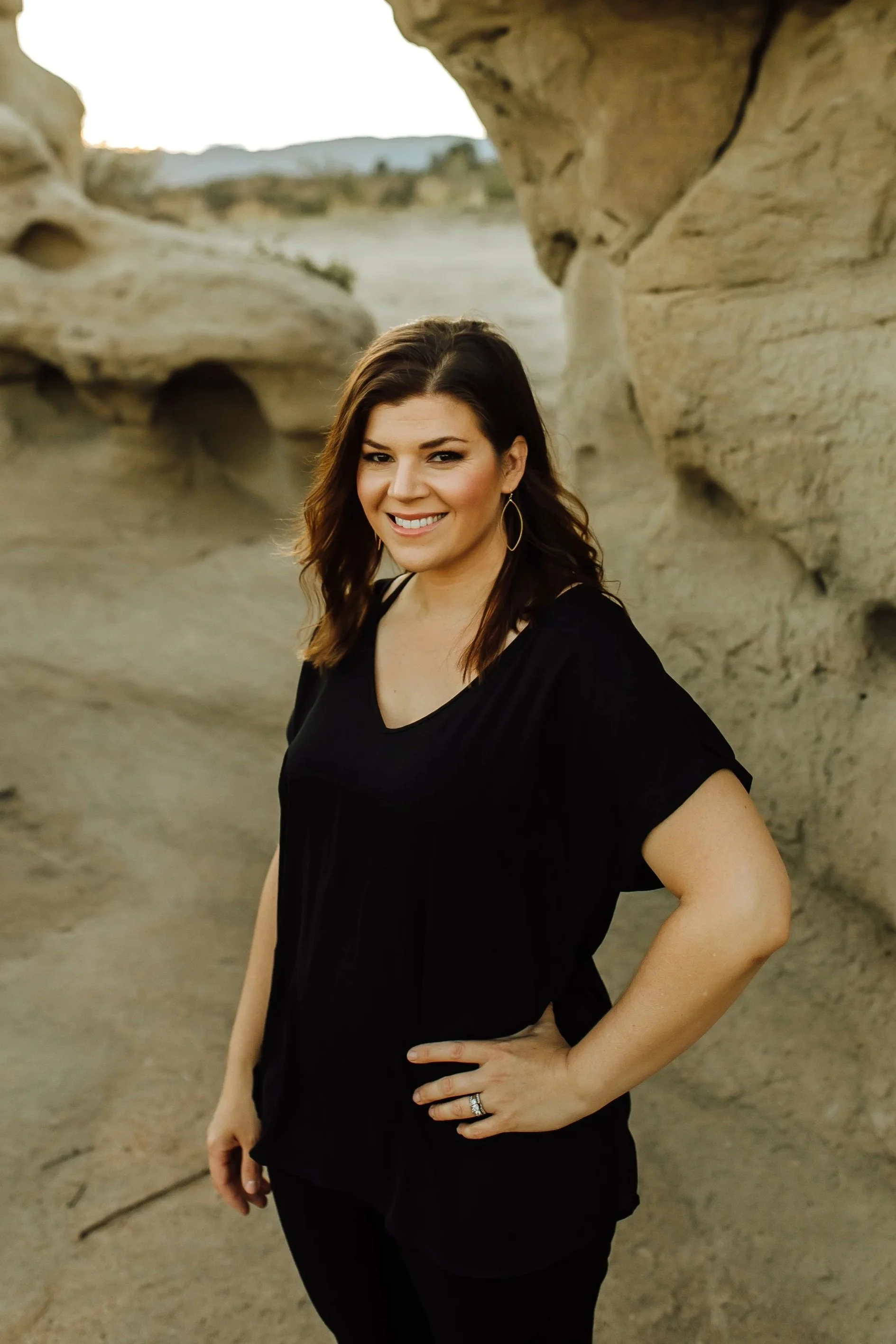 A woman with dark brown hair wearing a black shirt, standing outdoors in a rocky area with large sandstone formations and a blurred background of distant hills or mountains during sunset.