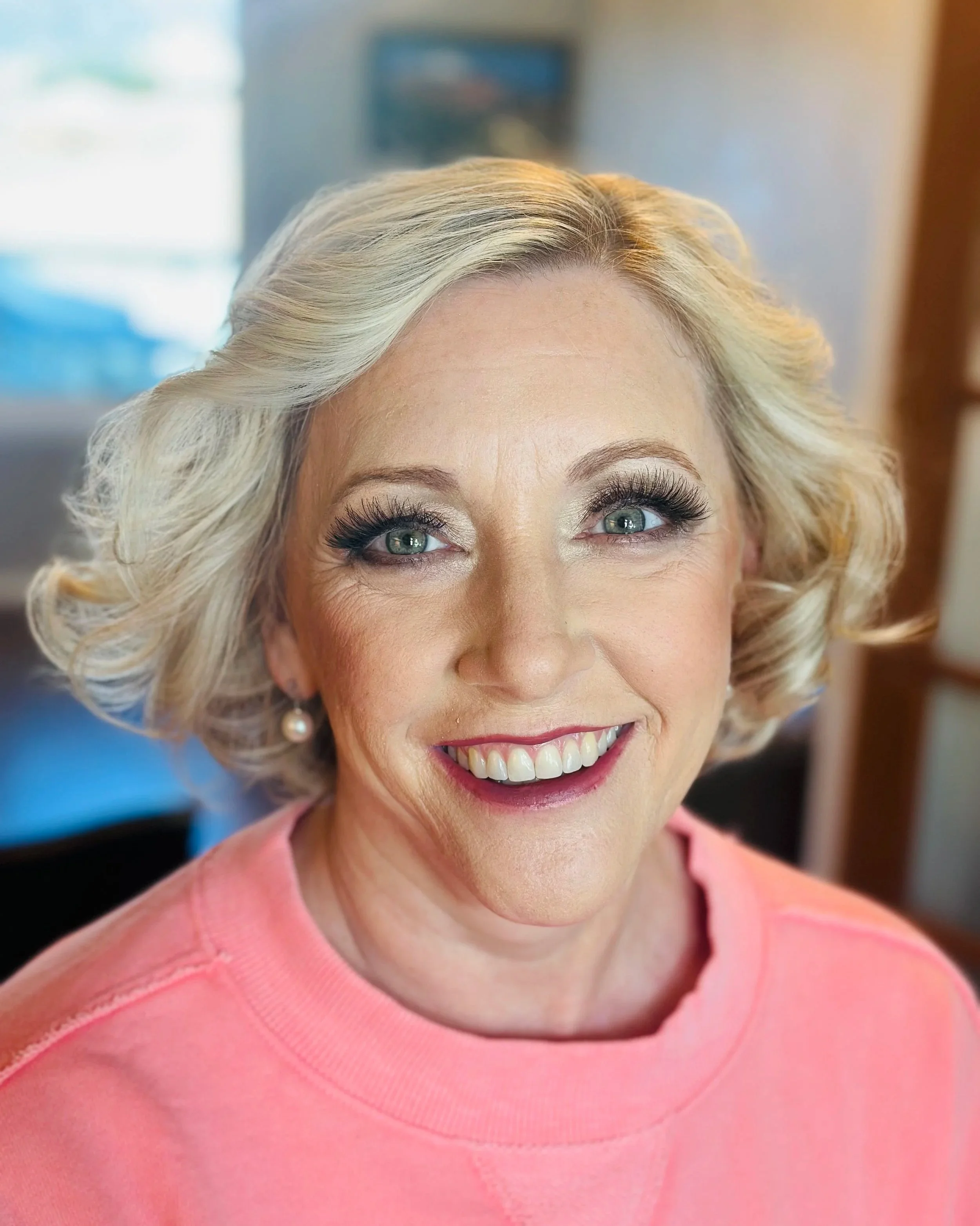 Close-up of a smiling woman with blonde curly hair, wearing makeup and a pink top, indoors.