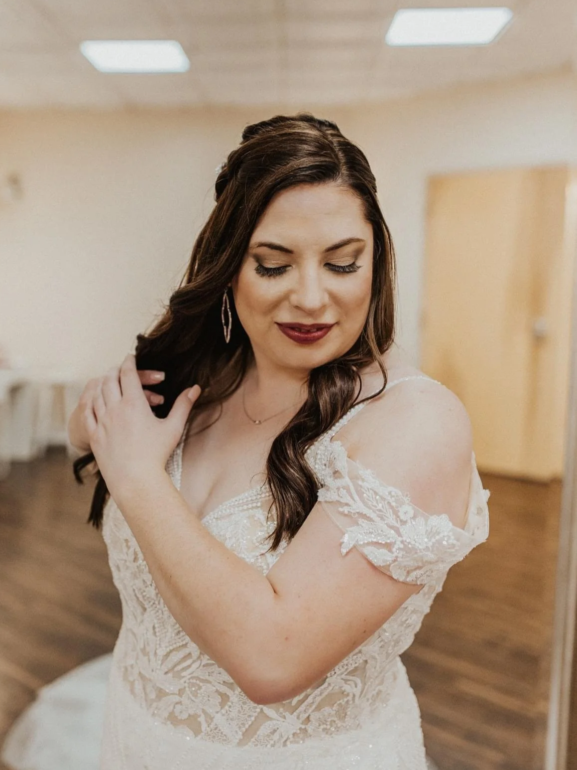 A bride with dark brown, wavy hair and makeup, wearing a lace wedding dress, is smiling with eyes closed and touching her hair in a softly lit room with wooden floor and light-colored walls.