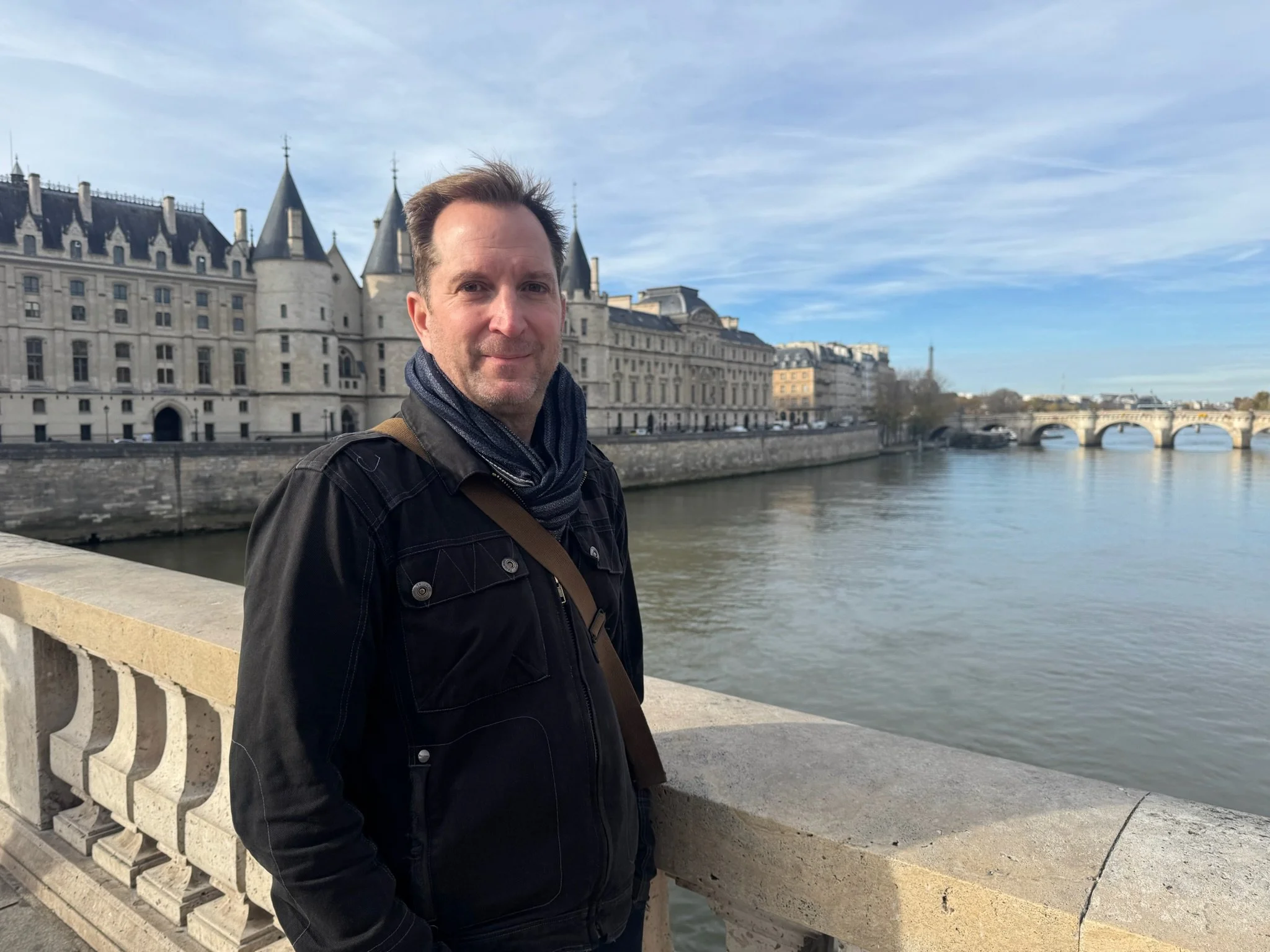 A man standing on a Paris bridge with historic buildings and the Seine River in the background, overcast sky.