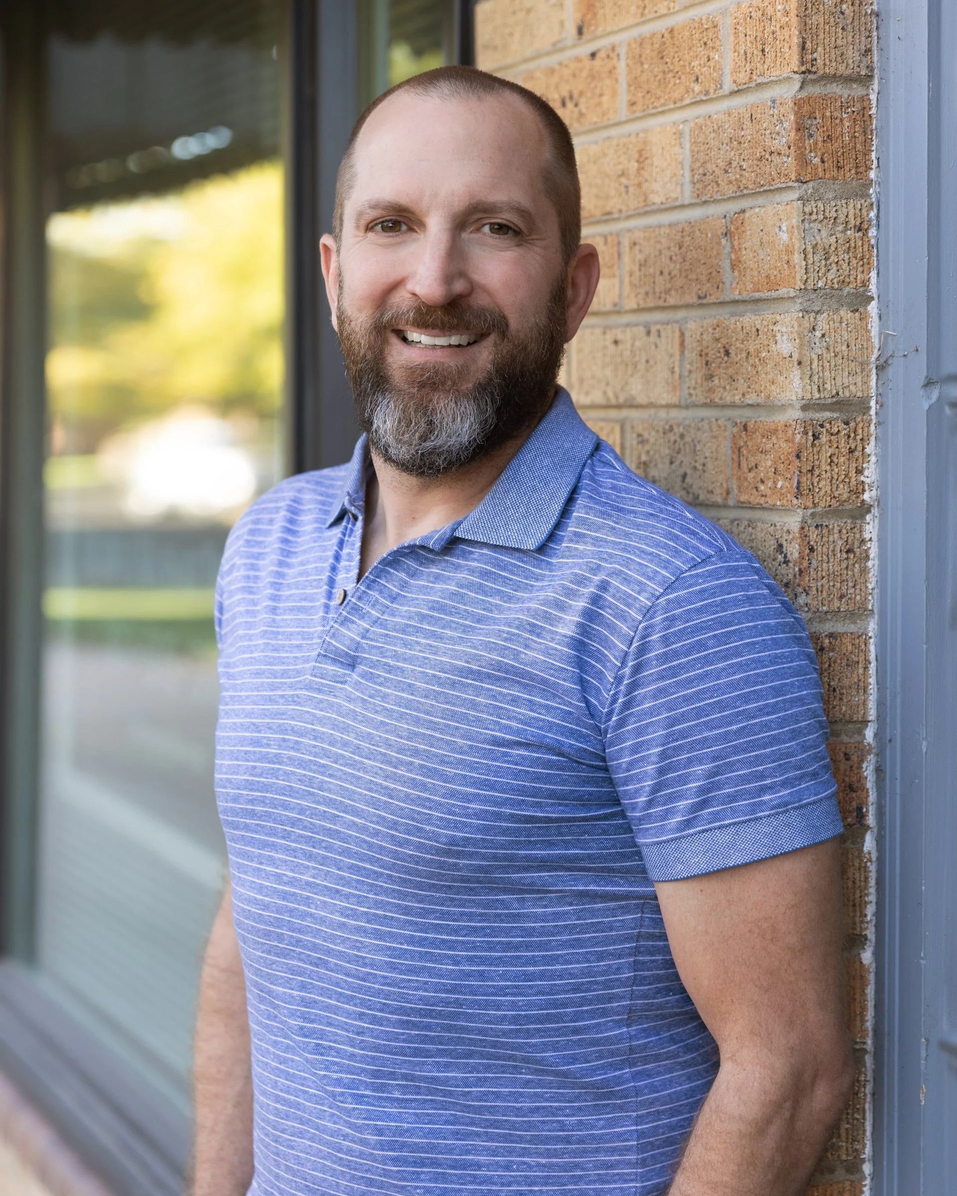 A smiling man with a beard and short hair standing outdoors near a brick wall, wearing a blue striped shirt, with a window and blurred greenery in the background.